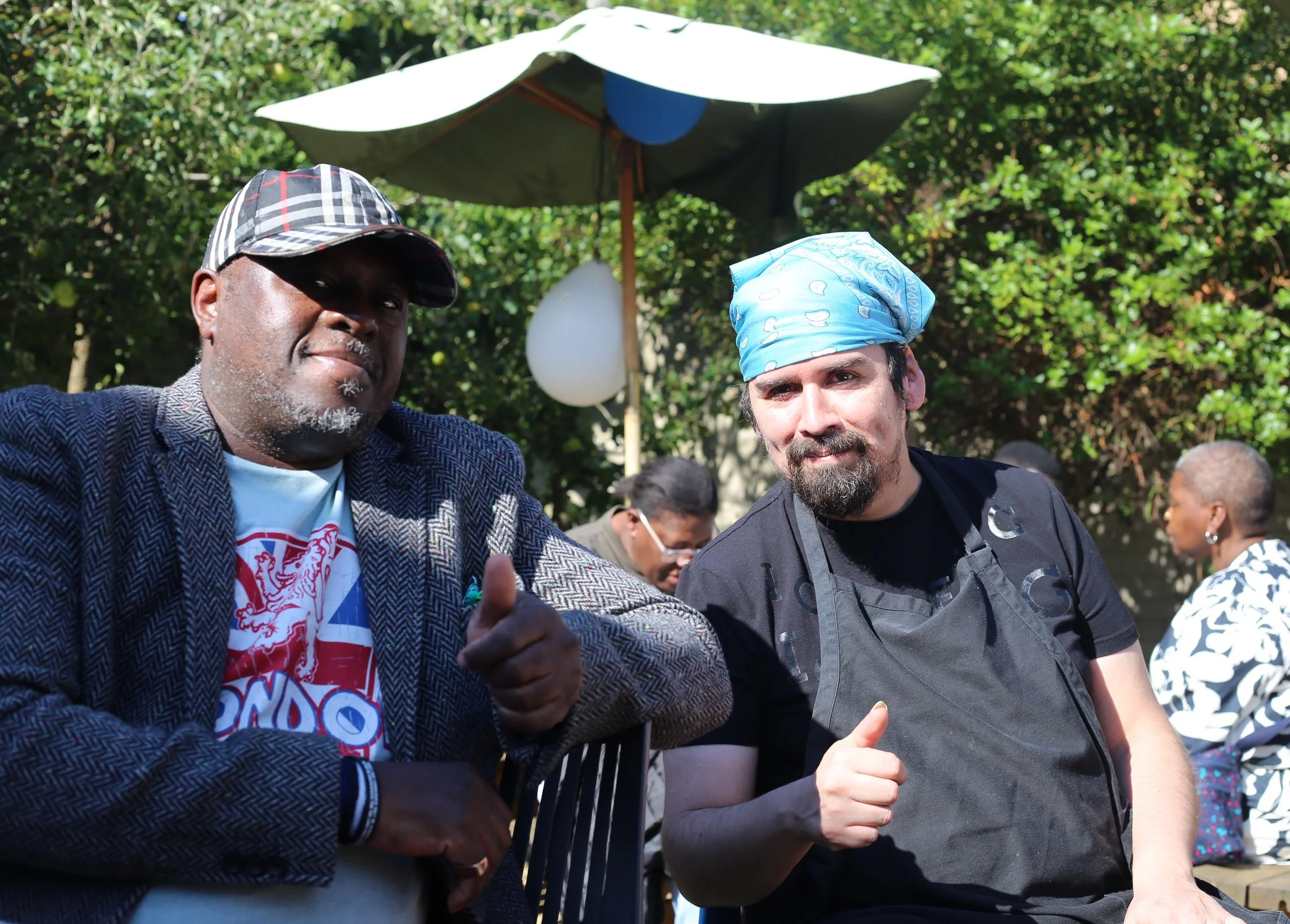 Two men at an outdoor event with greenery and a large umbrella in the background. One man is wearing a checkered hat and a blazer, and the other is wearing a blue bandana and a black t-shirt. They are posing for the camera, and the man with the blazer is giving a thumbs up.