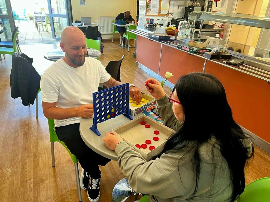 A man and a woman playing Connect 4 at a cafe table. The man is smiling, and the woman is holding a white flower. The cafe has colorful chairs, a counter in the background, and a person sitting further back.