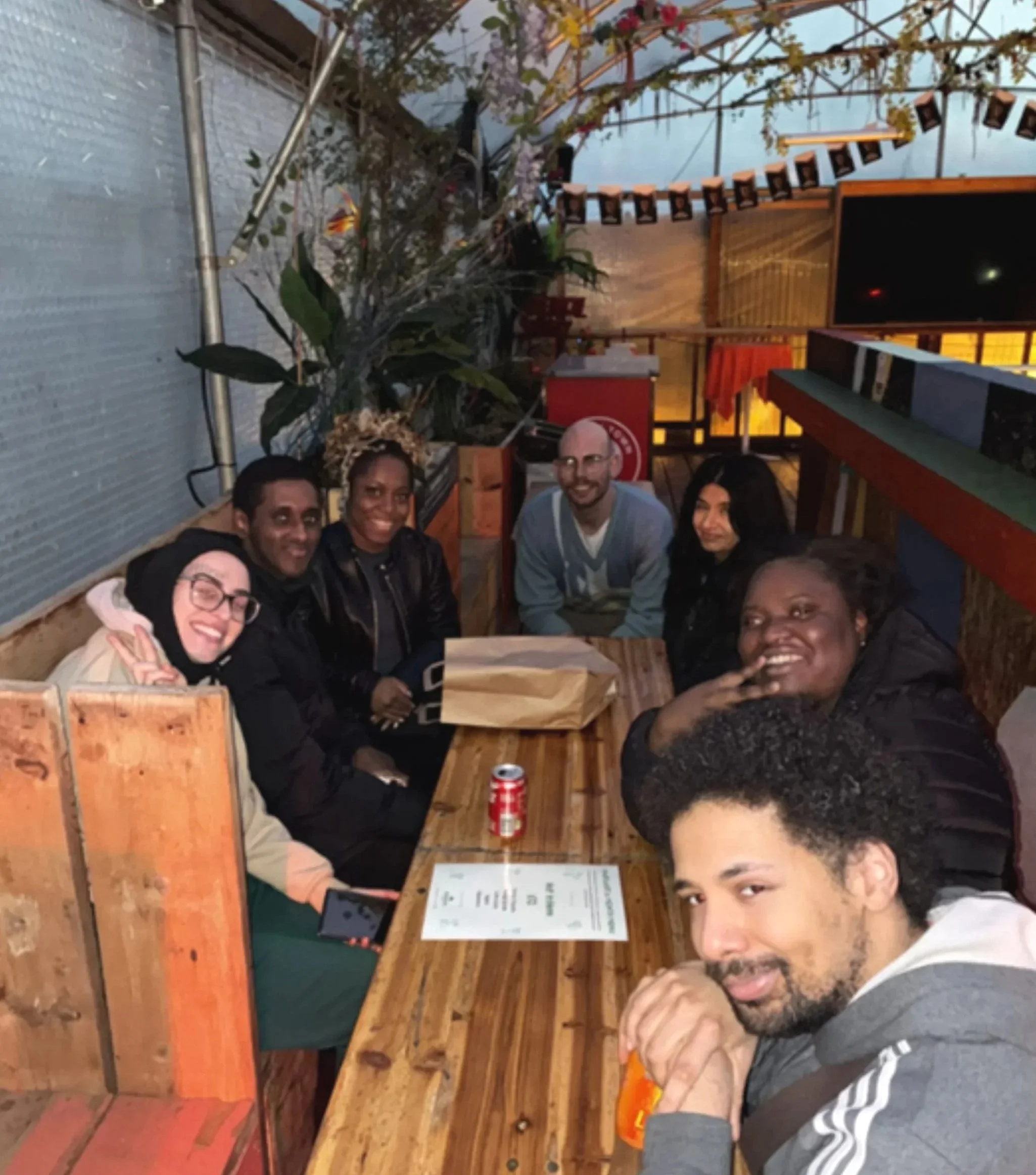 Group of diverse friends sitting at a wooden booth in a cozy indoor restaurant with wooden walls and string lights, smiling and enjoying their time together.