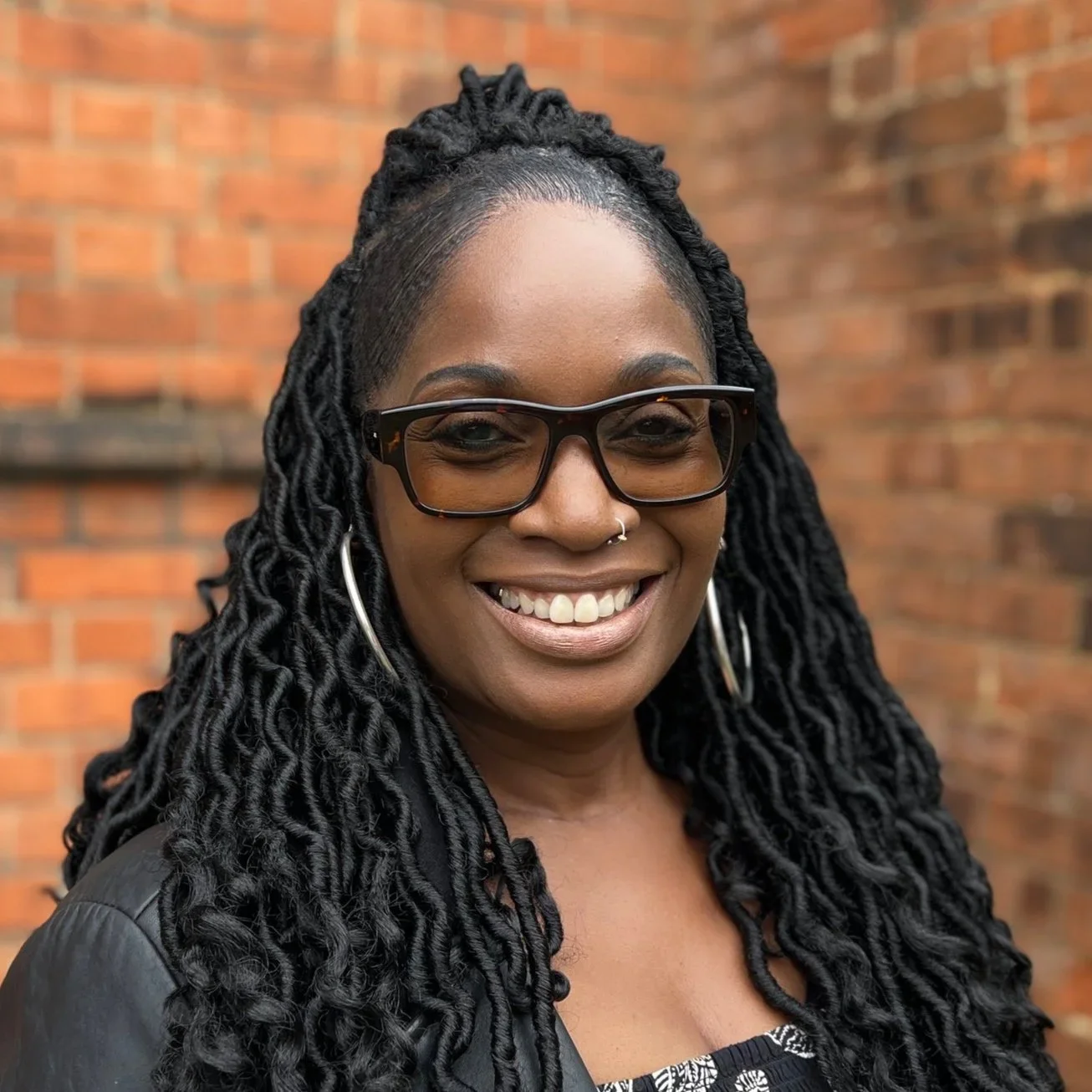 A smiling woman with long black dreadlocks, wearing glasses, hoop earrings, and a leather jacket, standing in front of a brick wall.