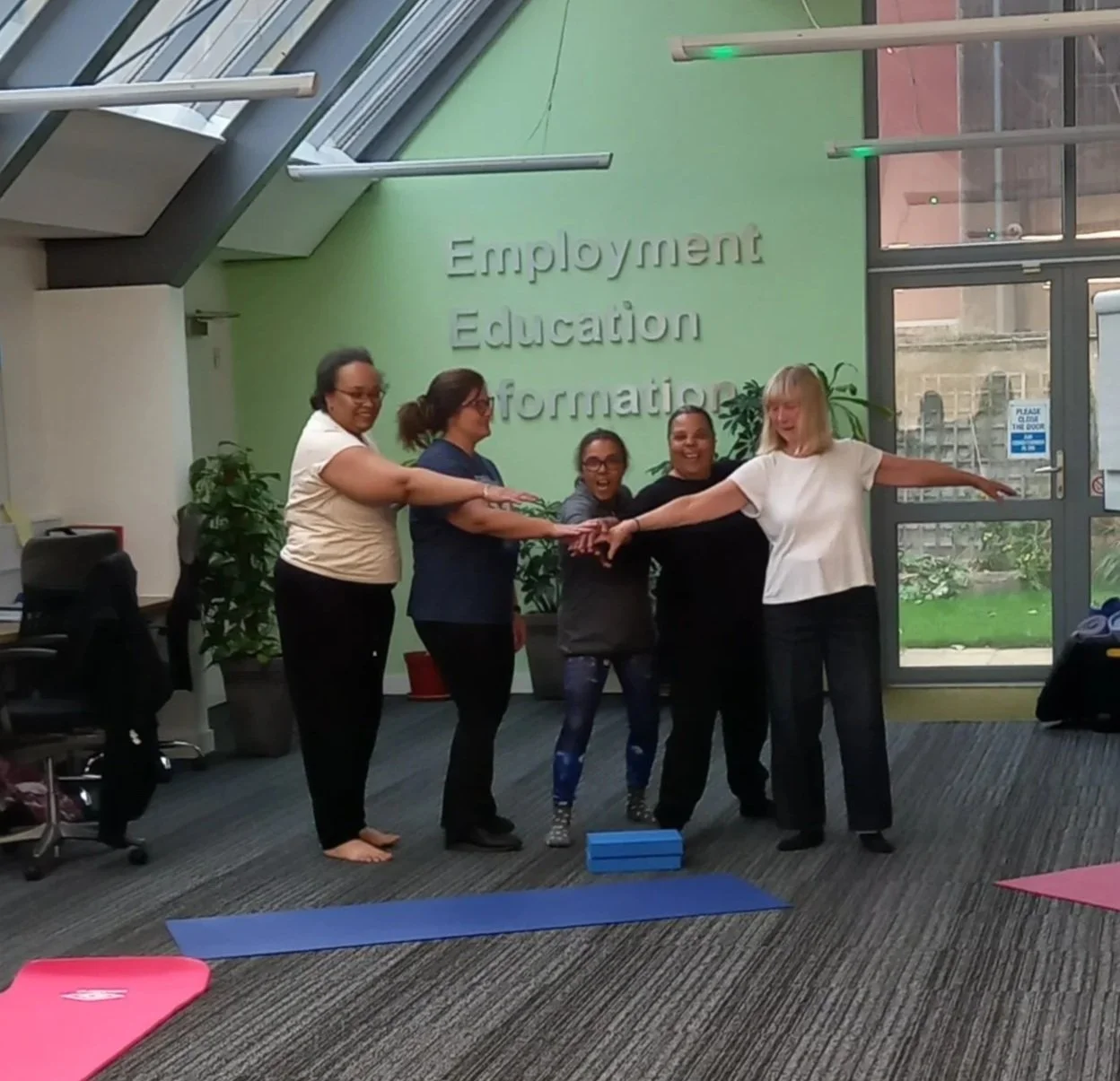 Five women standing inside a building, smiling, with their arms extended forward and touching hands. They are standing on mats, with yoga blocks on the floor. The background has a green wall with the words 'Employment Education Information.' Large windows let in natural light, and some plants and office furniture are visible.