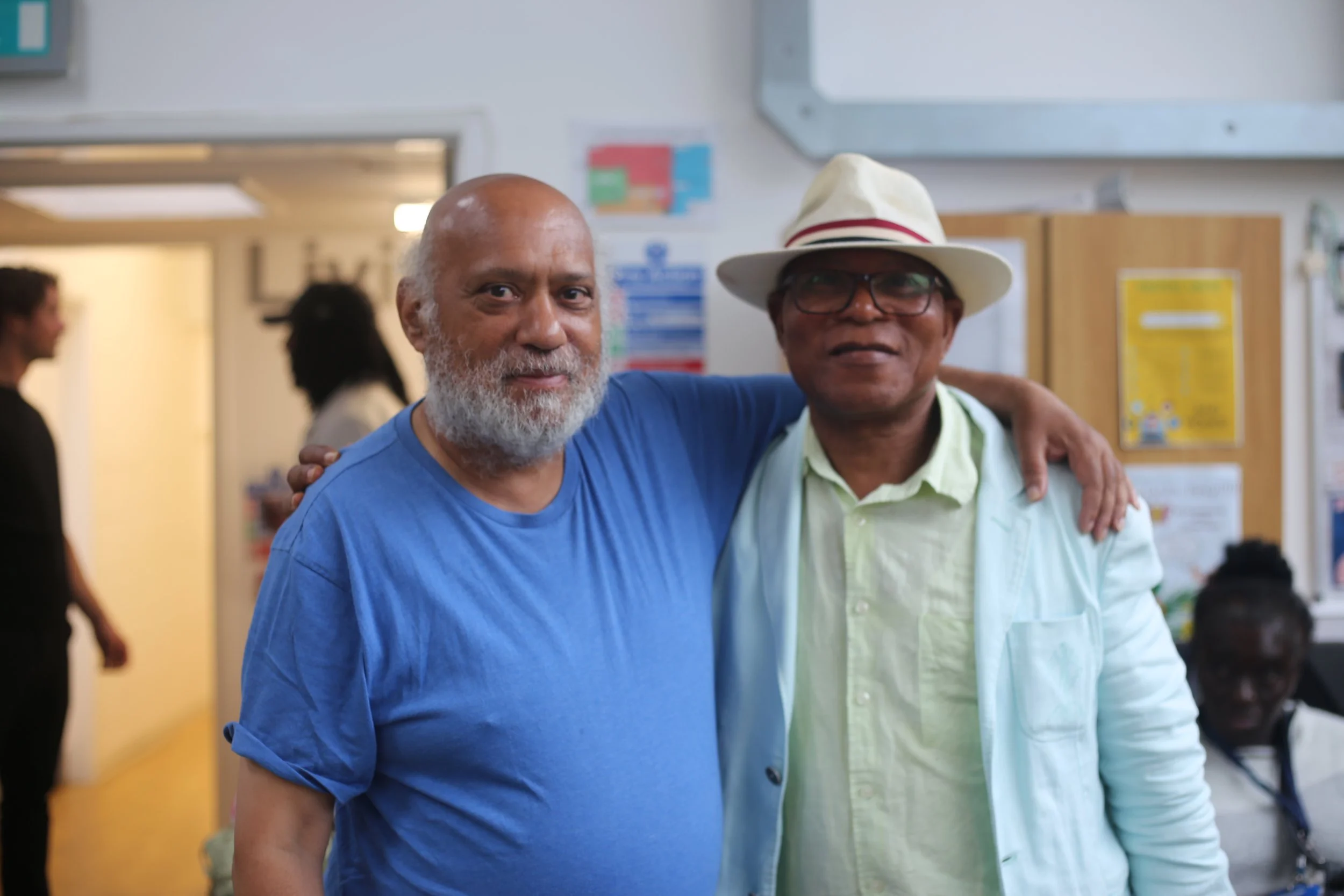 Two men, one with a beard wearing a blue shirt and the other wearing glasses and a white hat with a red band, are smiling and hugging inside a building with a yellow wall and colorful posters in the background.