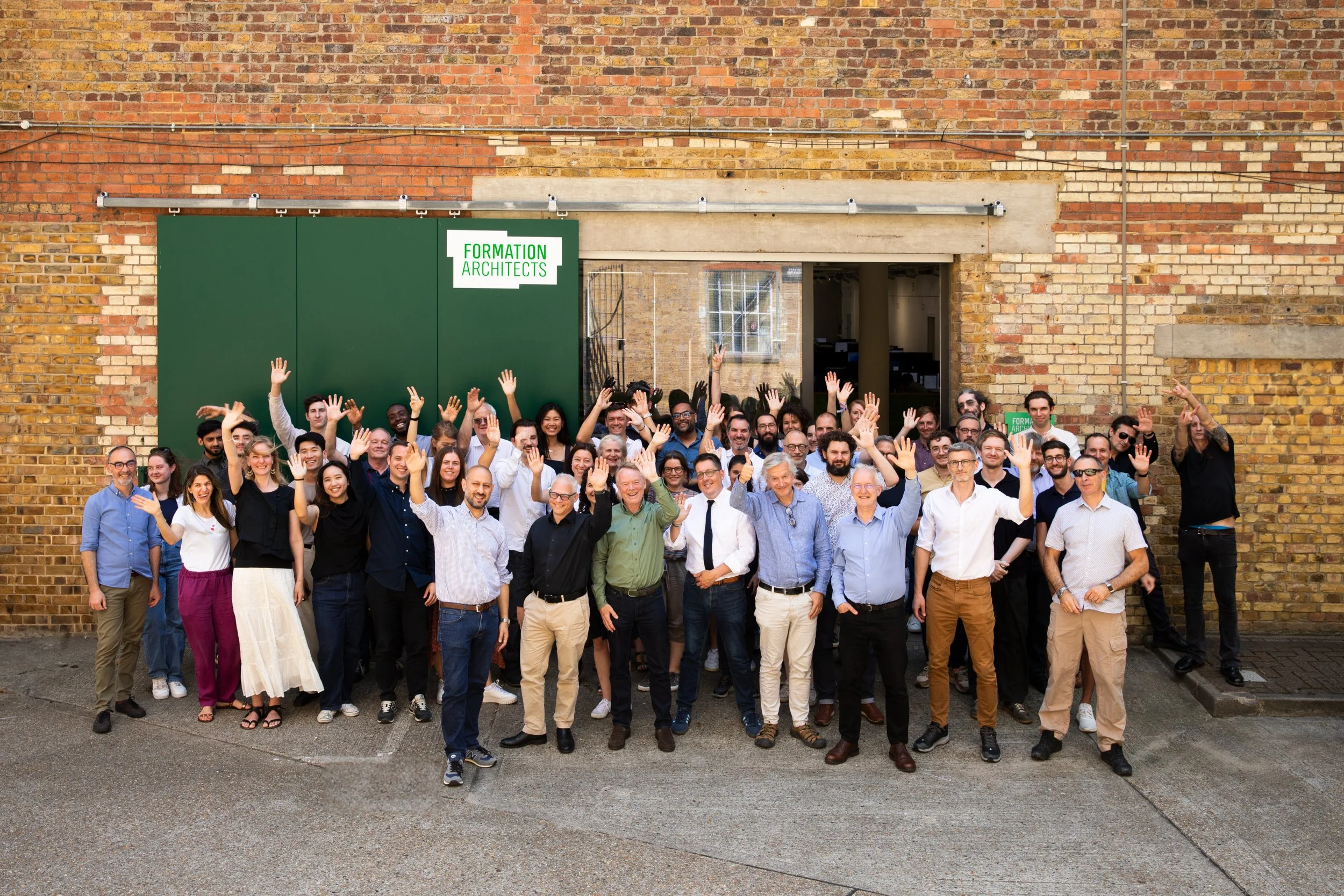 A large group of people standing outside in front of a brick wall, waving at the camera, with a sign that reads 'FORMATION ARCHITECTS' in green letters.