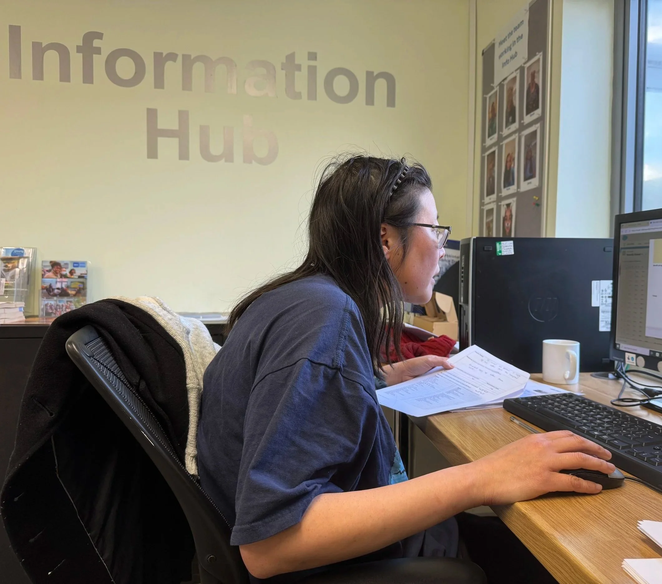 A woman with dark hair and glasses sitting at a desk using a computer, reading a document, with an information desk sign and photos on the wall behind her.