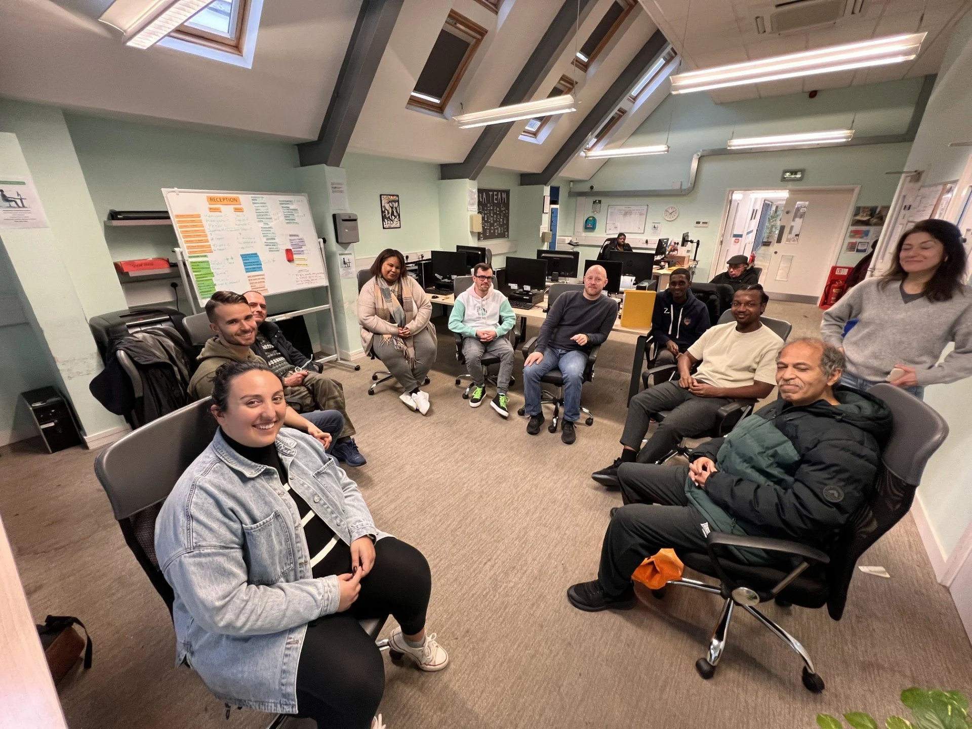 A diverse group of people sitting in a circle in an office or community room with computers, a whiteboard, and skylights.