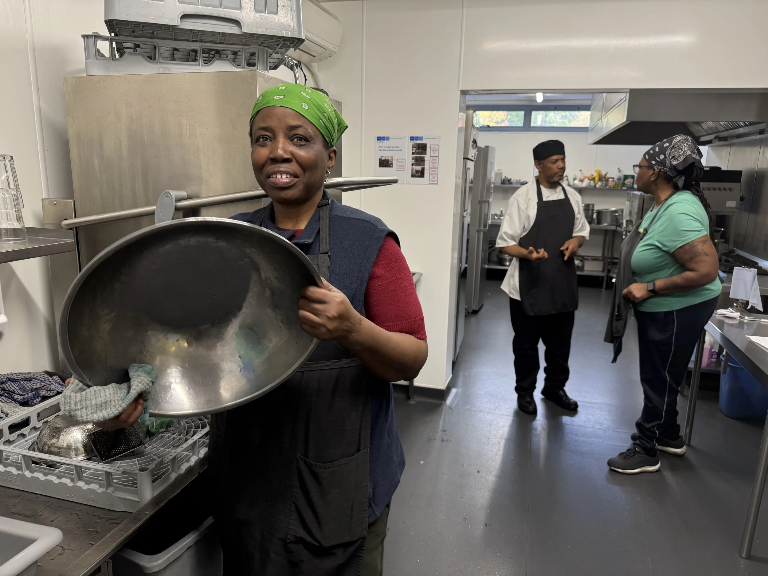 Three women working in a commercial kitchen, one in the foreground holding a large pot and wiping it with a cloth, the other two women in the background are talking and wearing aprons and head coverings.