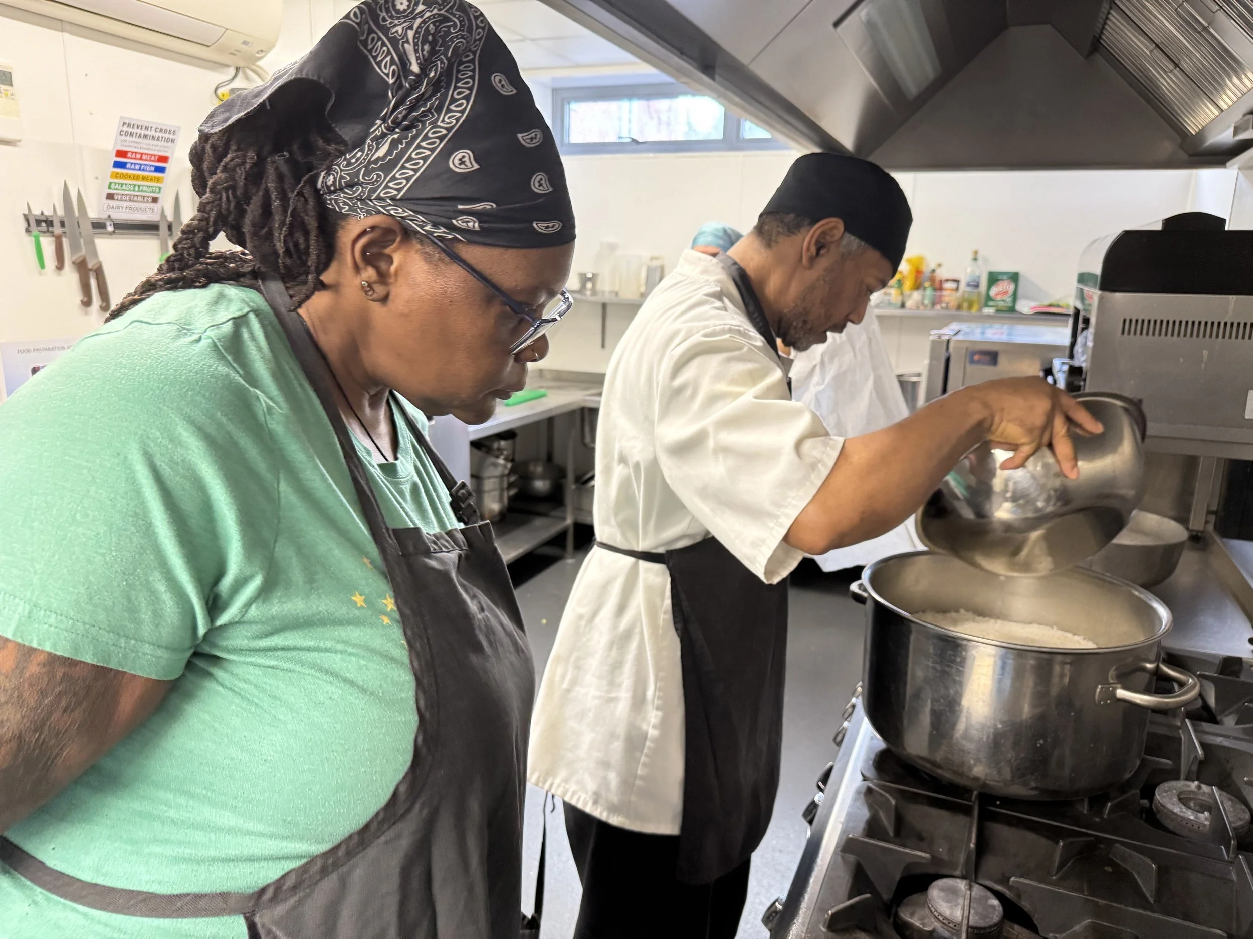 Two chefs working in a commercial kitchen, one woman with glasses and a bandana, and one man wearing a chef's coat and cap, both preparing food over large pots.