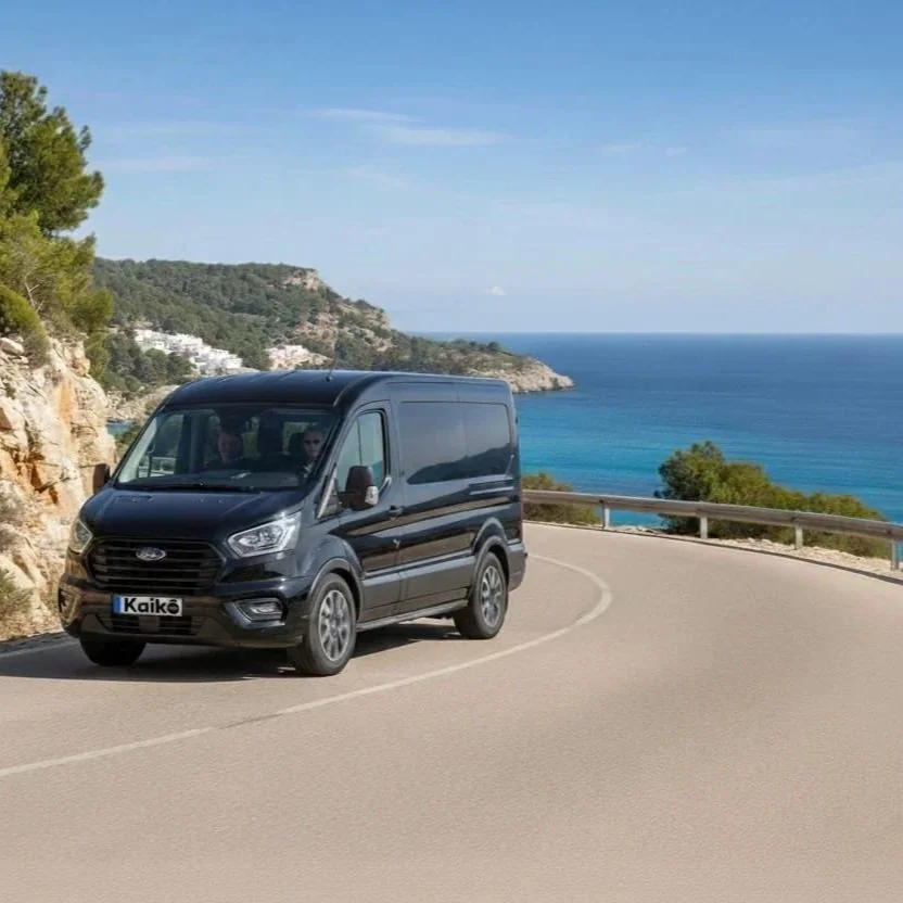 Una furgoneta negra conduciendo por una carretera costera con vista al mar y colinas con árboles al fondo.