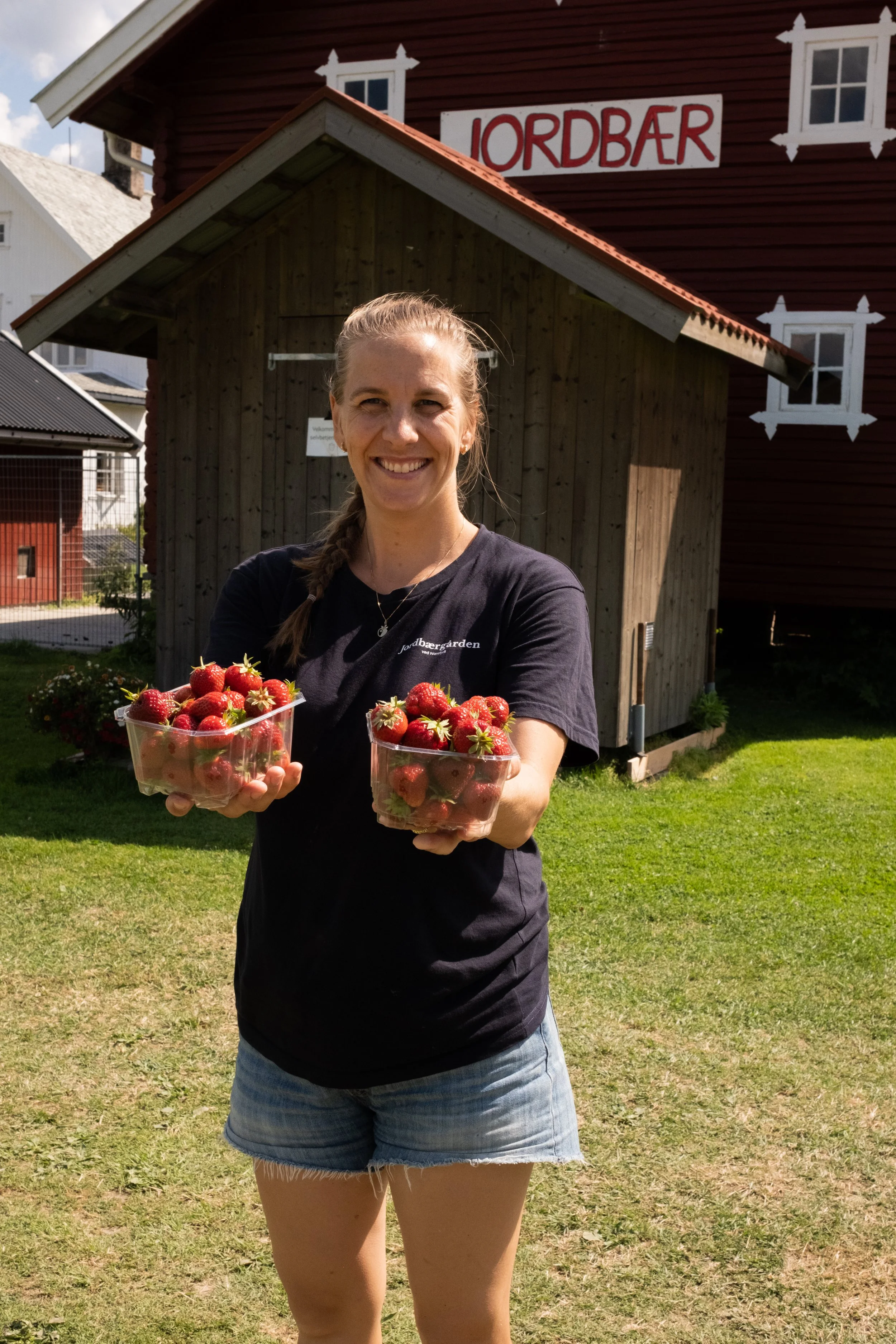 En kvinne smiler og holder to beholdere med jordbær foran en tradisjonell norsk bygning med skrift som sier 'Jordbær'.