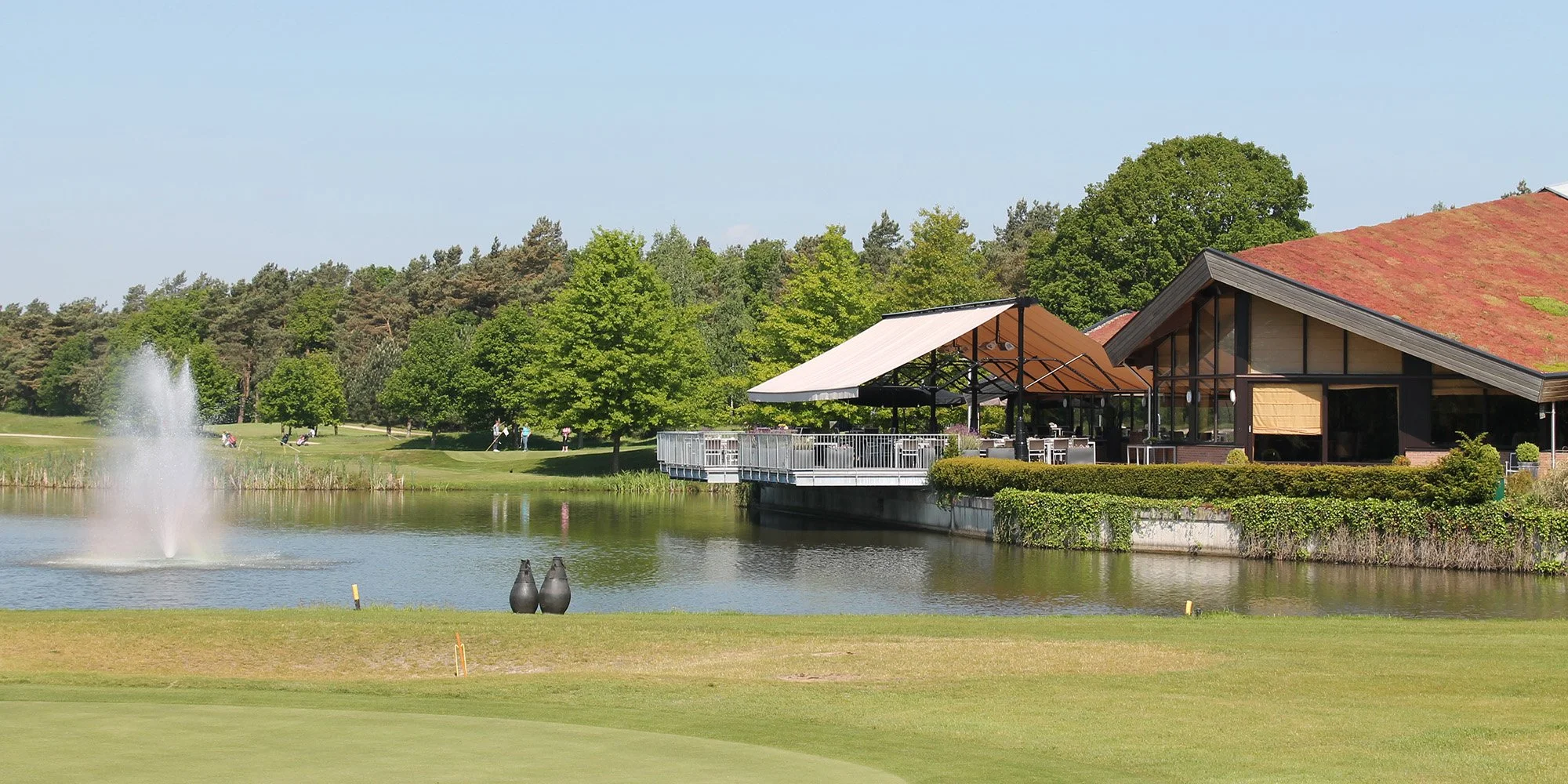 Een rustige golfbaan met een meer, een fontein, omgeven door groene bomen en een gebouw met grote ramen en een terras, onder een blauwe hemel.