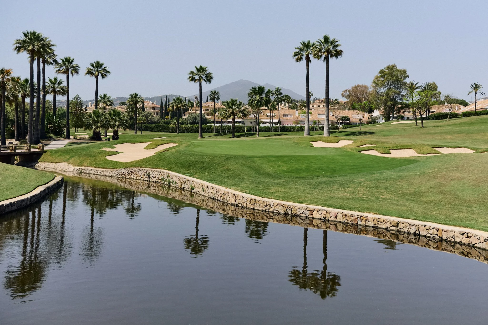 Golfbaan met palmbomen en een waterpartij, huizen in de achtergrond en bergen op de horizon.