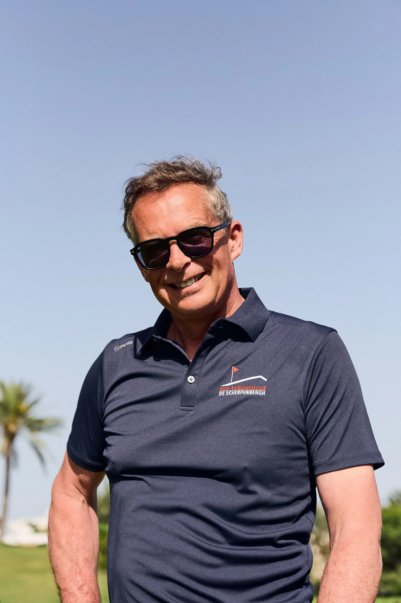 Man met grijs haar en zonnebril, lachend, in een donkerblauw polo met logo, buiten op een zonnige dag.