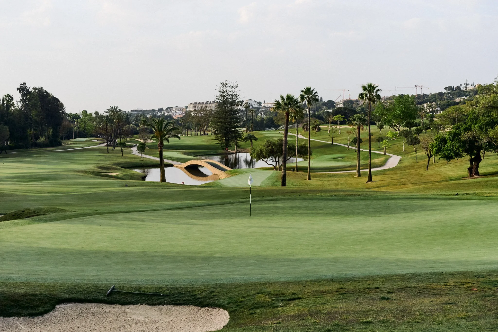 Een golfbaan met een puttinggreen, waterpartij en palmbomen, in de verte gebouwen en bouwkranen te zien.