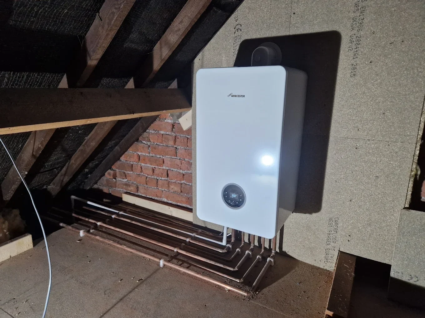 A white Worcester boiler mounted on a wall with pipes beneath, in an unfinished room with brick and wood framing.