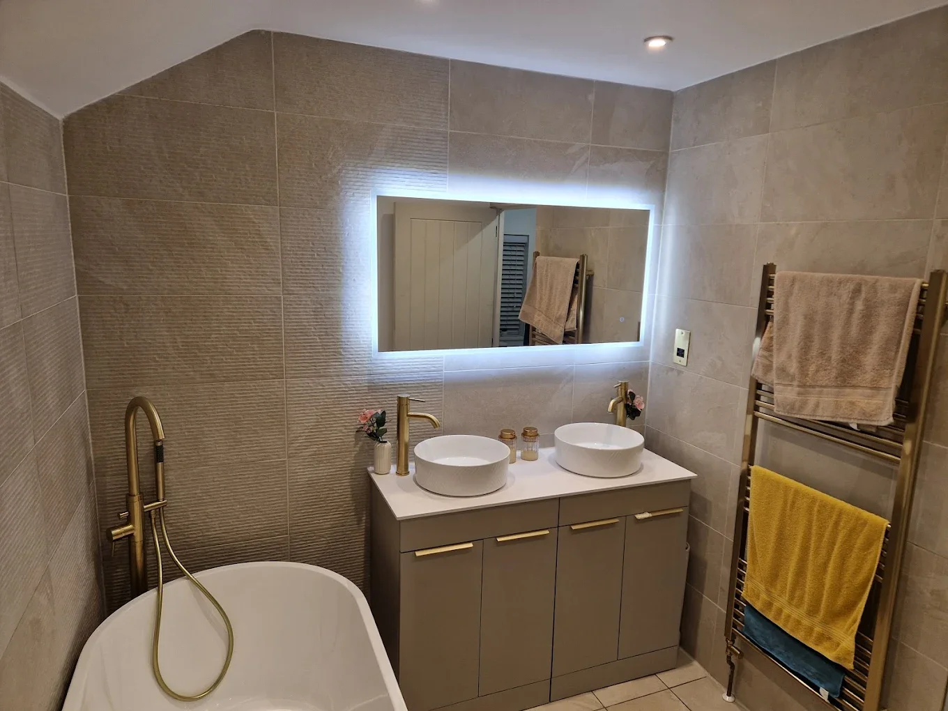 Modern bathroom with beige tiled walls, double vessel sinks with gold faucets, illuminated mirror, bathtub with gold fixtures, beige towels on a gold towel warmer, and small flower decorations on the countertop.