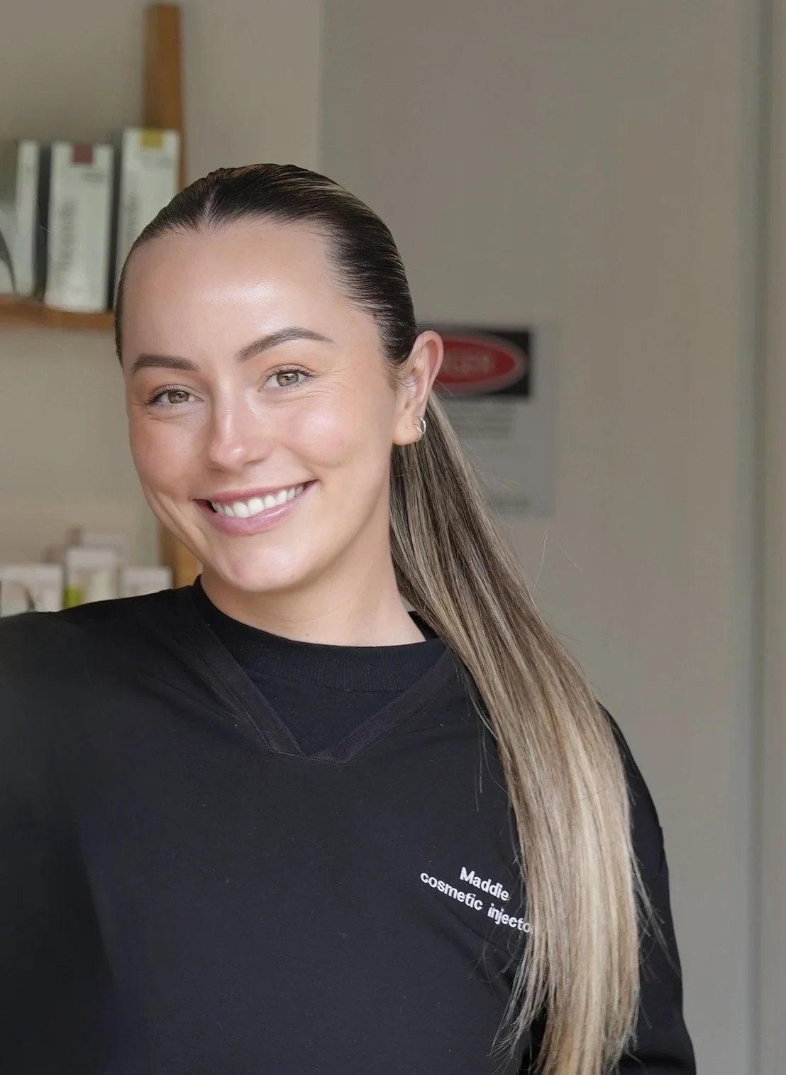 Smiling woman with long hair and earrings wearing a black shirt that says 'Maddie cosmetic injector' in a room with shelves of products in the background.