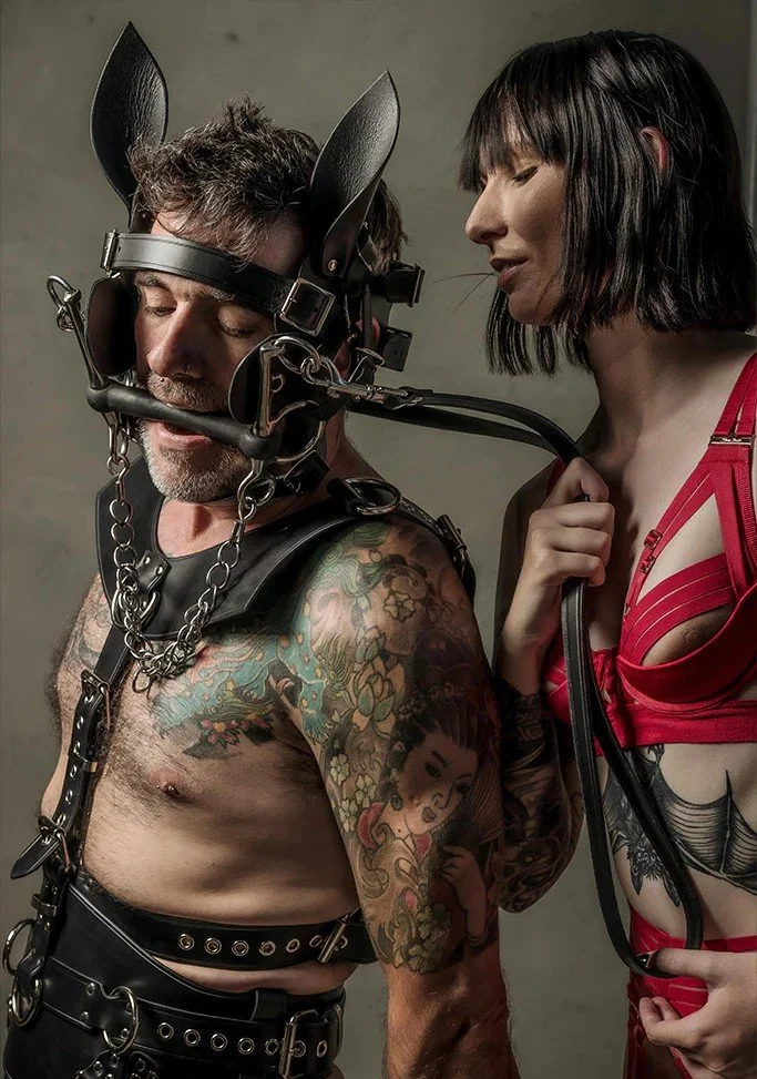 A man with tattoos on his arm wearing BDSM gear, including a leather harness and a headgear with bunny ears, is being held by a woman dressed in red lingerie, holding a leash attached to his headgear.