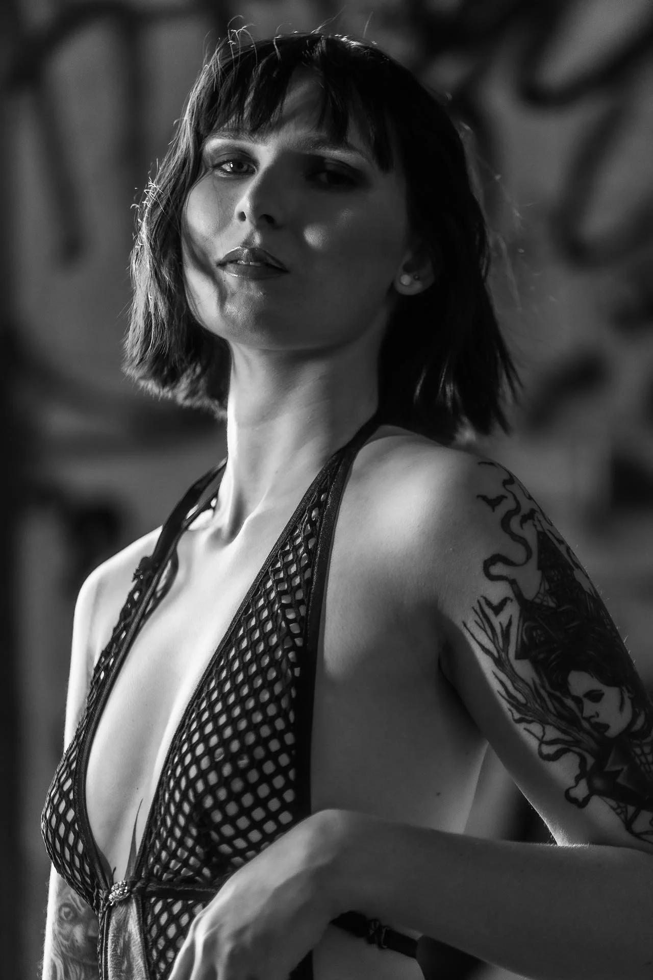 A black and white photo of a woman with short hair, tattoos on her arm, wearing a fishnet top with a zipper, and looking slightly to the side with a confident expression.