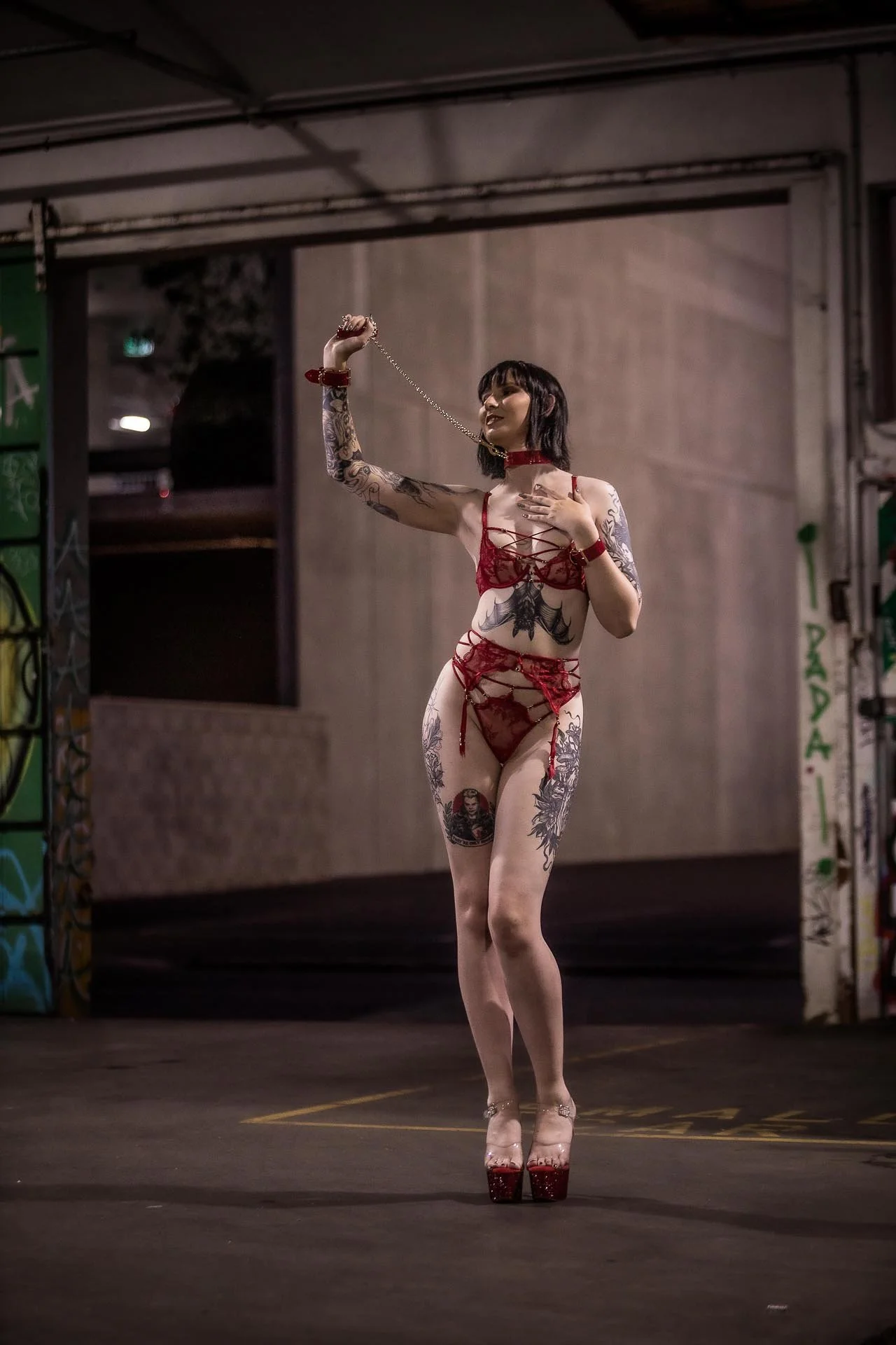 A woman with tattoos in red lingerie and high heels stands in an urban setting, holding a chain near her face.