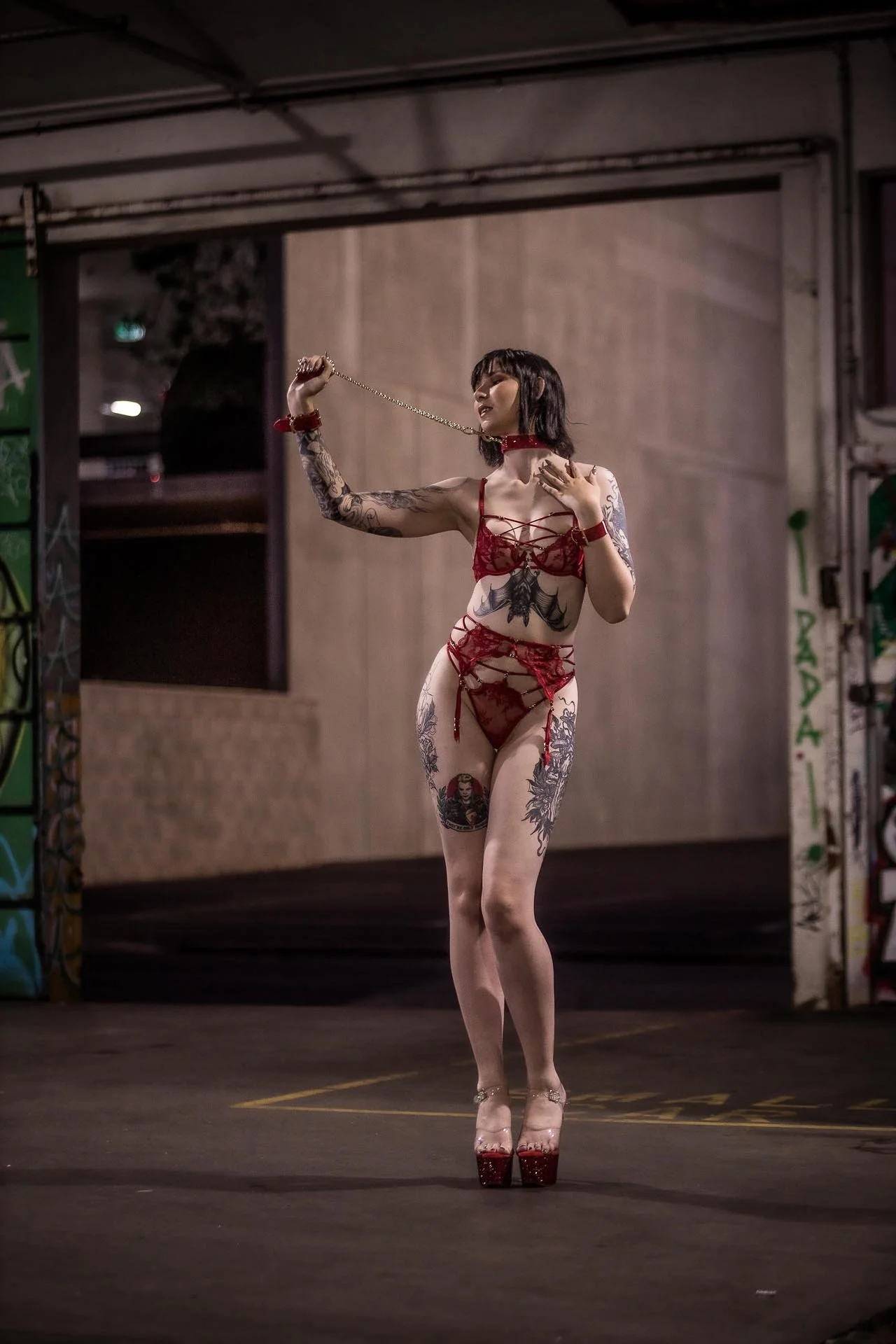 A woman in red lingerie and high heels stands in an urban, graffiti-covered setting, posing with a chain in her hand.