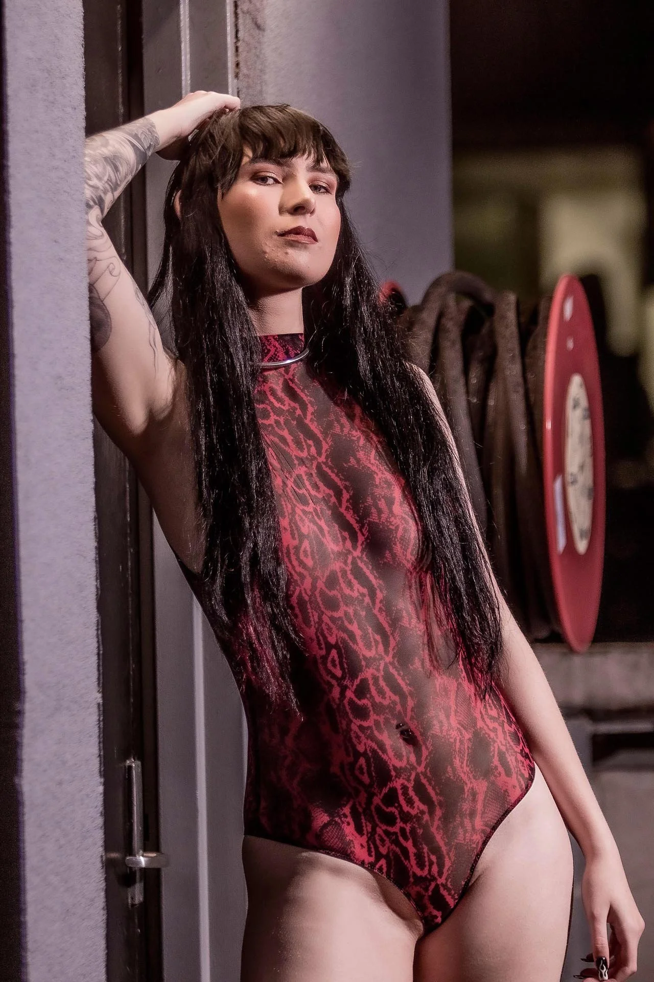 A woman with long dark hair, wearing a sheer red and black leopard print bodysuit, standing against a wall with her left hand resting on her head and looking at the camera.