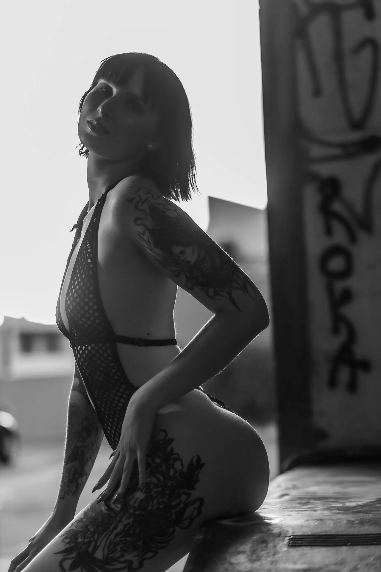 A woman with tattoos posing on a ledge in black and white, wearing a revealing mesh outfit.