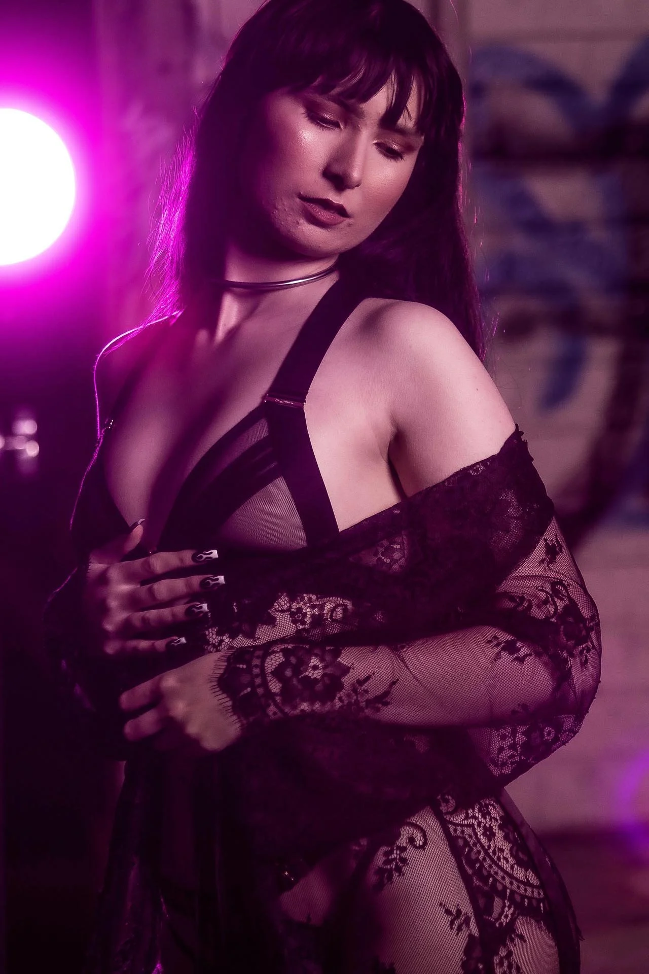 A woman with dark hair and makeup wearing black lingerie and lace gloves, posed with her eyes closed in a dimly lit setting with purple lighting.