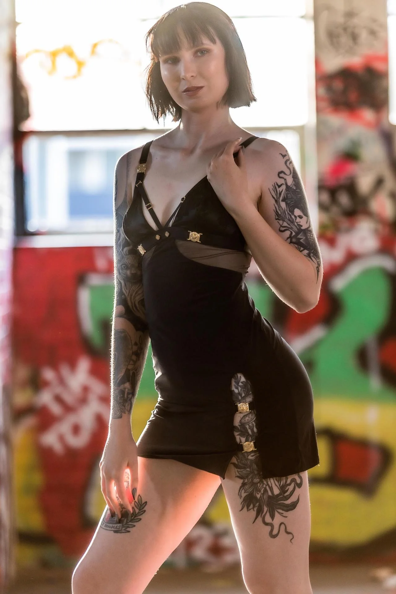 A woman with short dark hair and tattoos wearing a black dress with gold accents, standing against a graffiti-covered wall in front of a sunlit window.