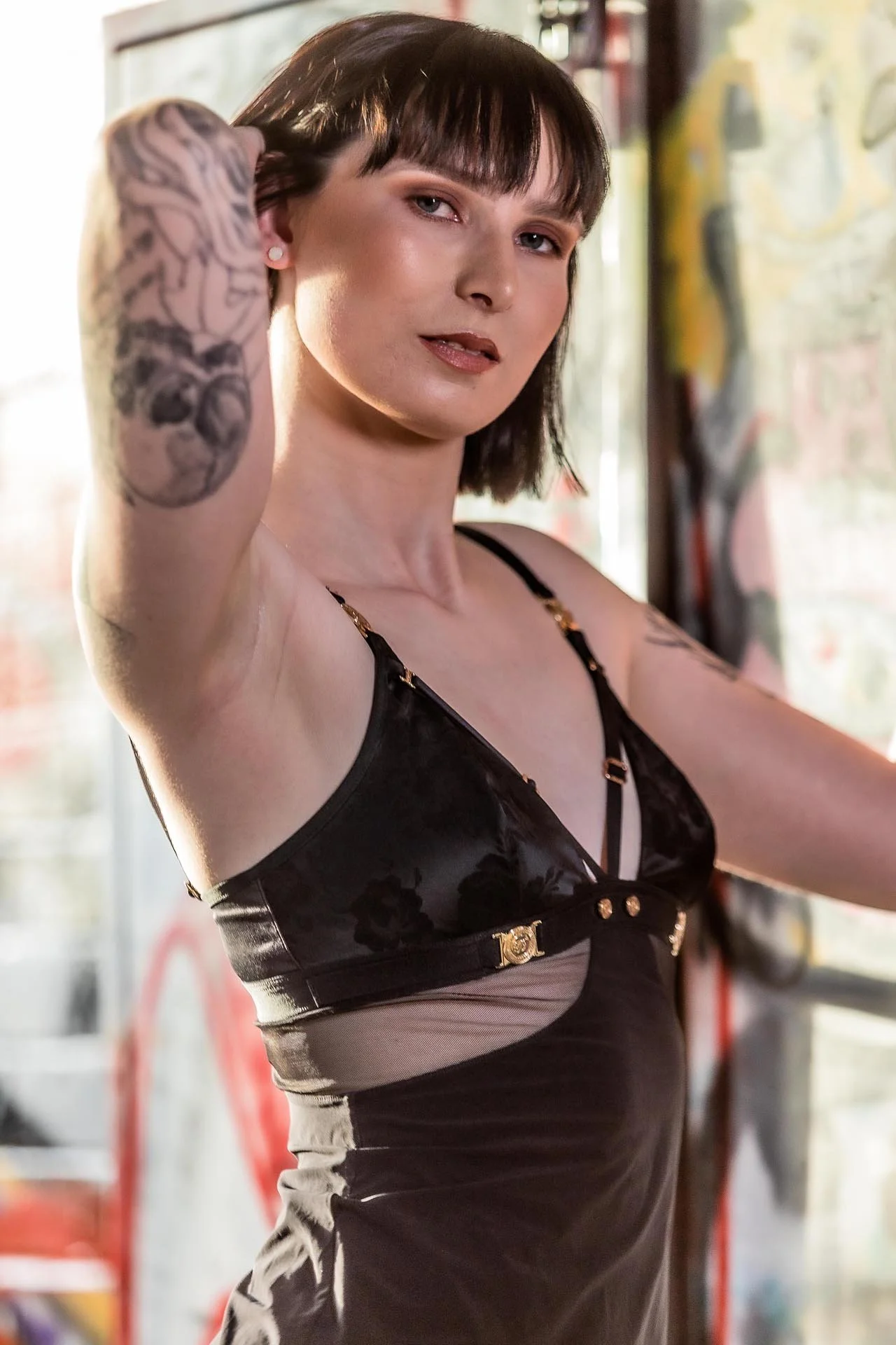 A woman with short dark hair and tattoos on her arms, wearing a black lingerie top, posing indoors with sunlight coming through the window.