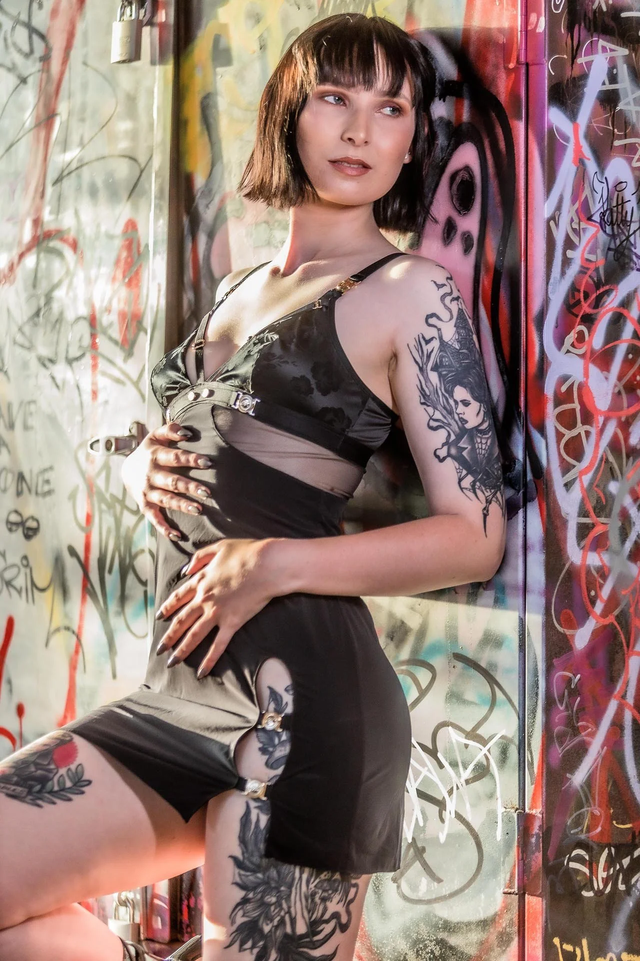 A young woman with short dark hair and multiple tattoos, wearing a black dress, standing against a wall covered in colorful graffiti.