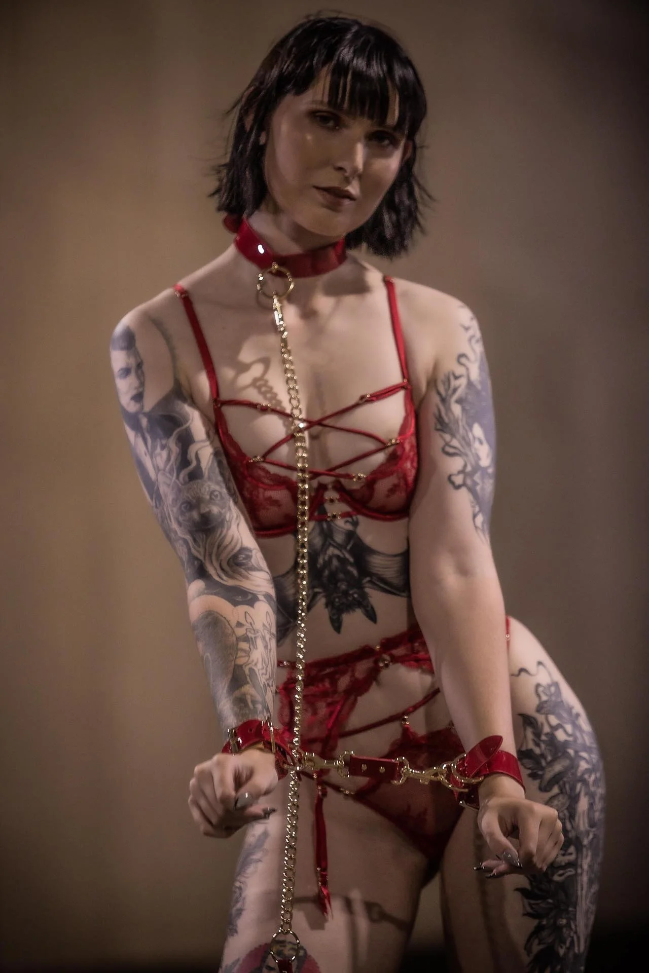 A woman with dark, torn short hair and tattoos on her arms and torso, wearing red lingerie with various straps, cuffs, and chains.
