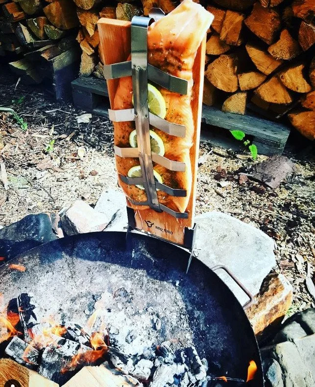 A cut of salmon with lemon slices held upright over a campfire with wood logs in the background.