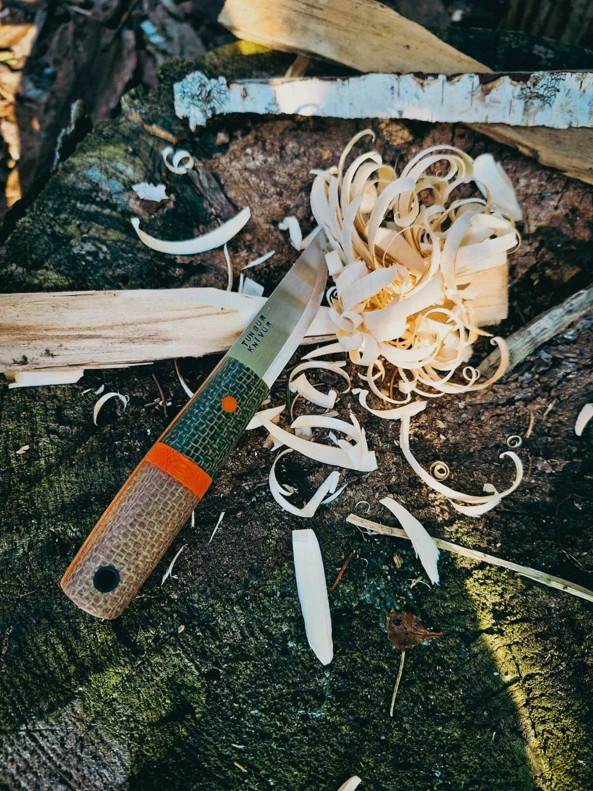 A woodworking knife with a textured handle, cutting into a piece of wood on a mossy tree stump, producing wood shavings.
