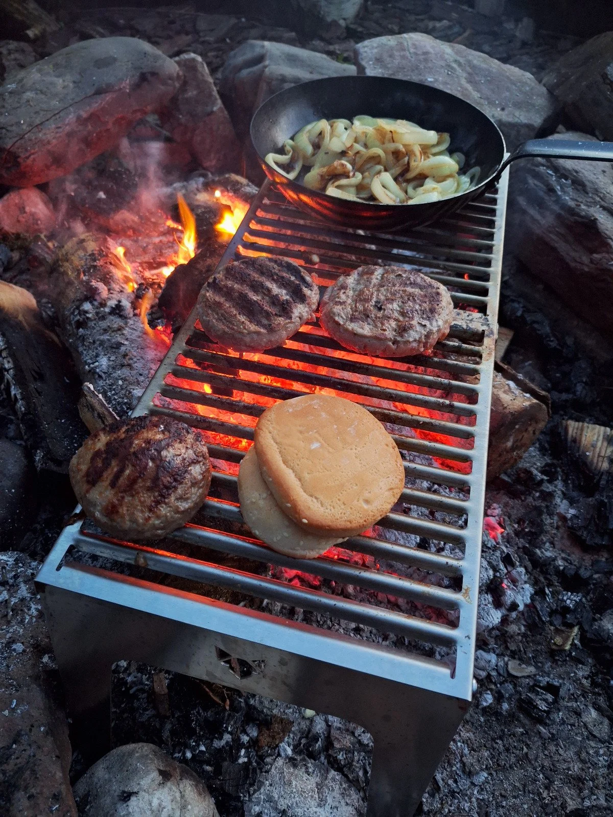 Hamburgers cooking on a grill over an open fire with a bun on one patty, and a bowl of sautéed onions nearby.