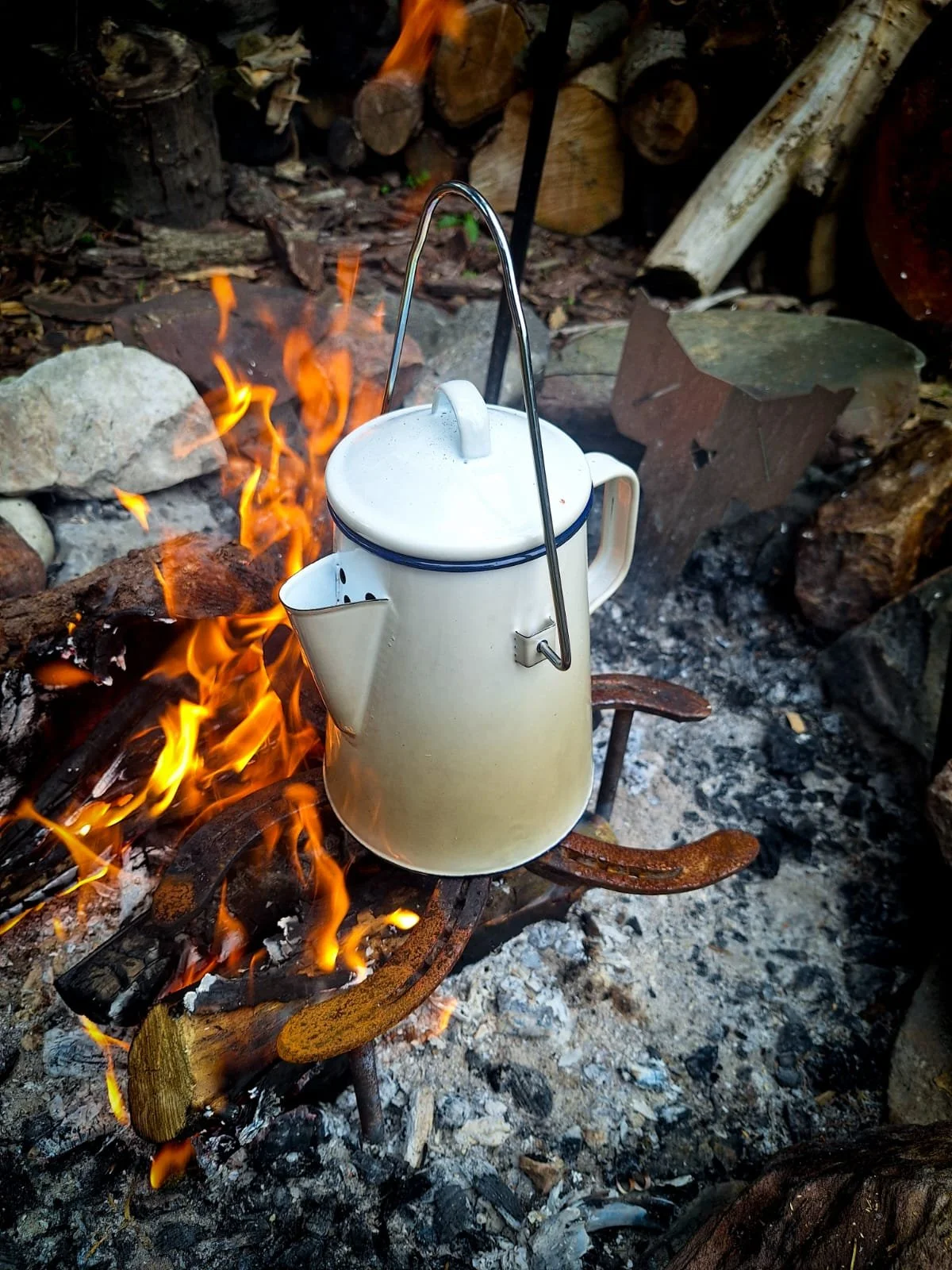 Enamel kettle hanging over an open campfire with wood logs in the background.