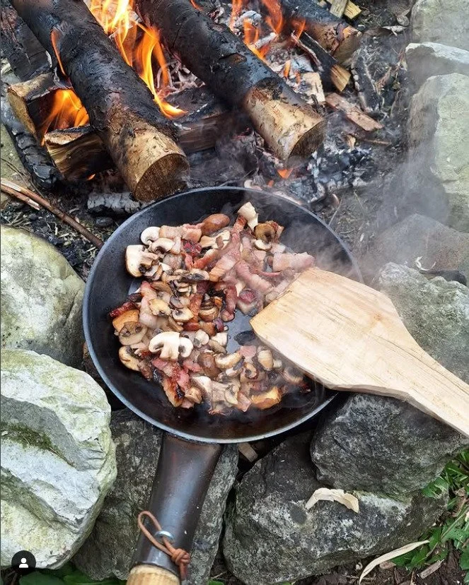 Cooking mushrooms and bacon in a black frying pan over an outdoor campfire surrounded by rocks.