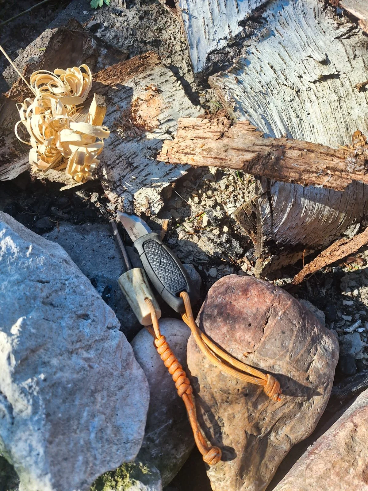 A pocket knife, rocks, and wood shavings surrounded by fallen wood and rocks, likely outdoors.