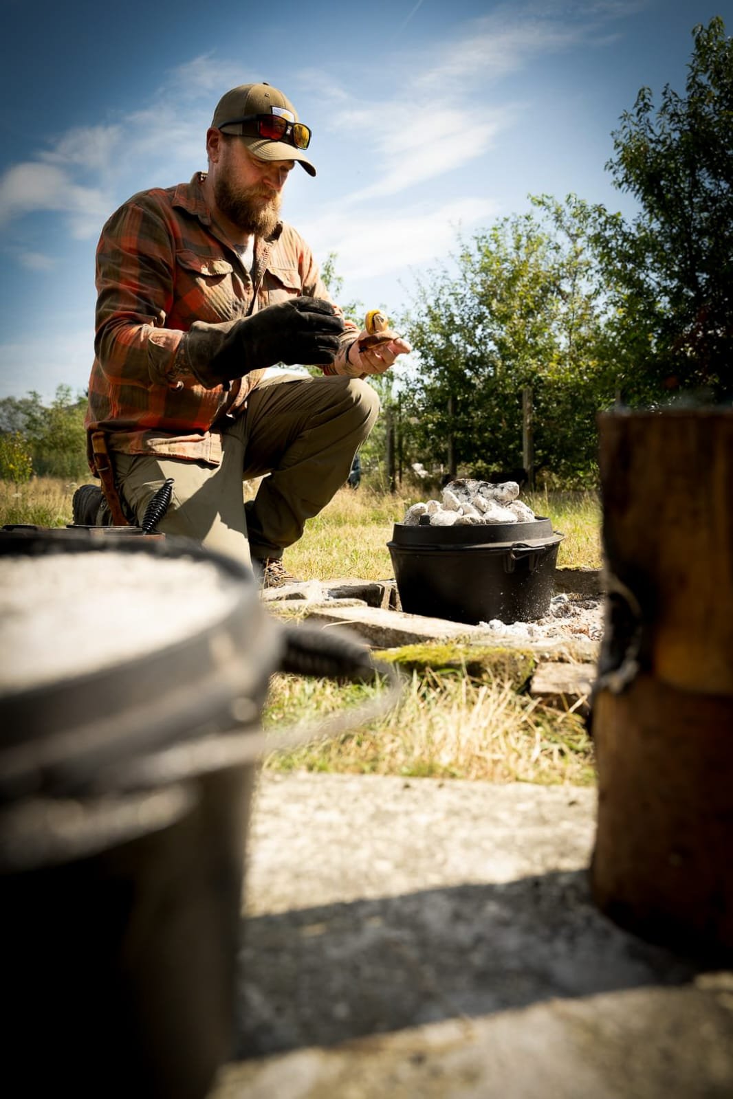 Man kneeling outdoors, examining a small object, with a fire pit containing rocks behind him and greenery around, under a partly cloudy sky.