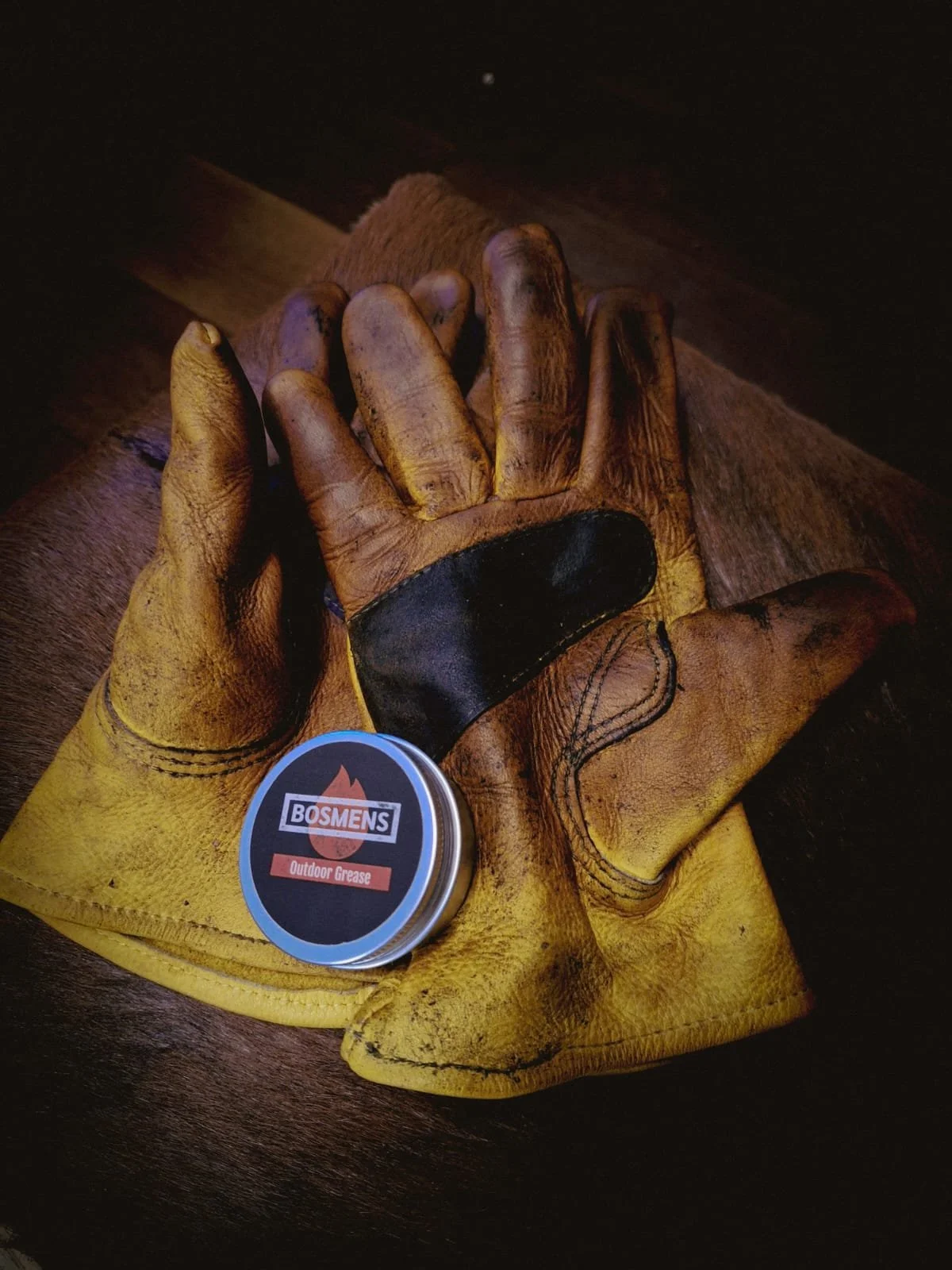 A pair of well-worn yellow work gloves with black padding on the palms, resting on a dark wooden surface. In front of the gloves is a small round container labeled "Bosmens" with "Outdoor Grease", indicating it is a lubricant for outdoor use.
