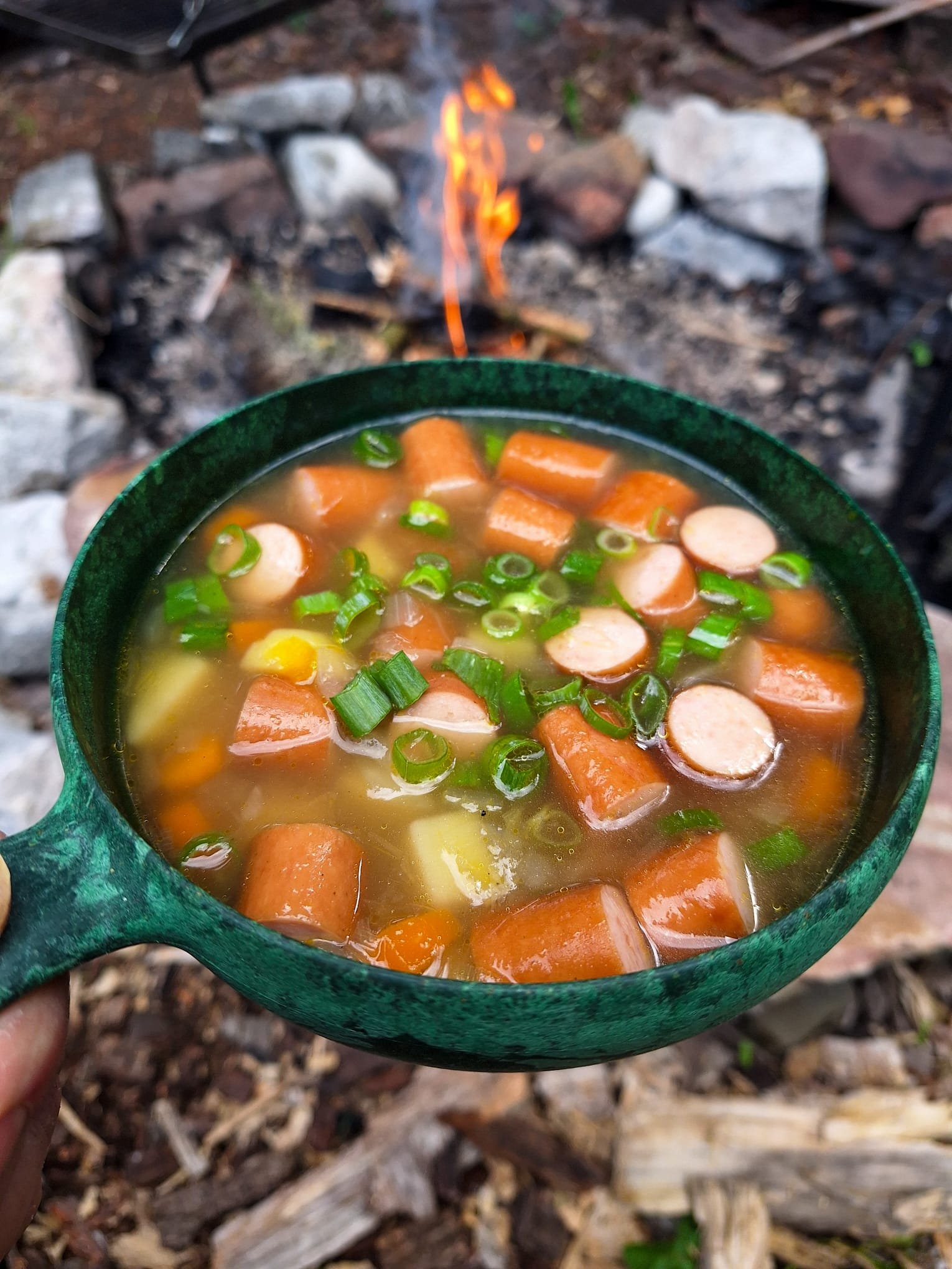 A green camping bowl filled with soup containing sliced sausage, carrots, potatoes, and green onions, held in front of a campfire with rocks around it.