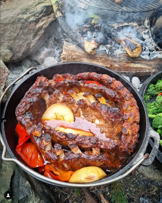 A cast iron skillet on an outdoor fire showcases a beef brisket roast glazed with sauce, garnished with roasted red bell peppers, onions, garlic cloves, and potato slices, with a smoky fire in the background and logs.