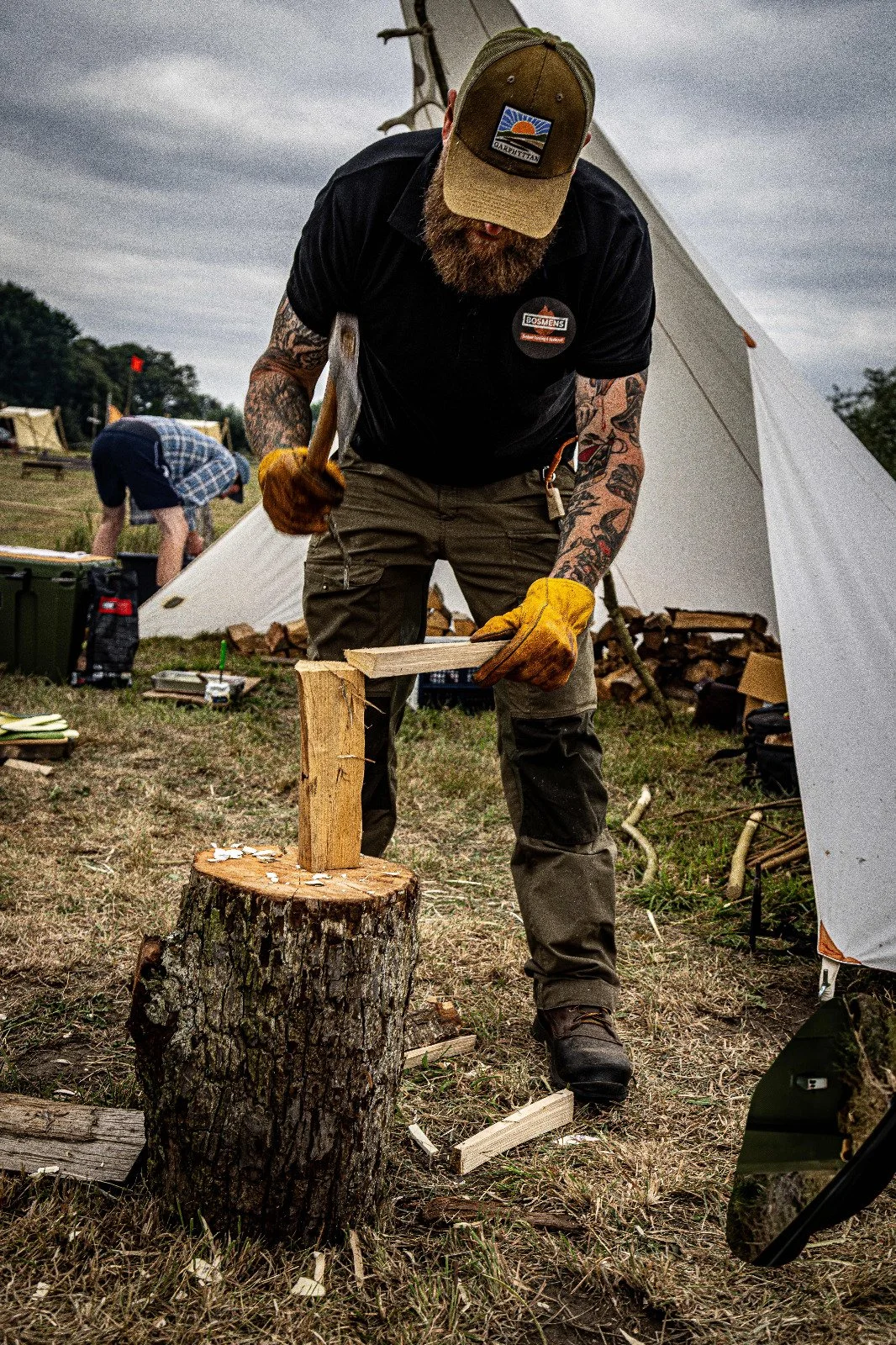 A man with a beard, wearing a cap, black shirt, and yellow gloves, is chopping wood with an axe. He is standing on the grass in front of a tree stump, with a white tent and another person in the background.