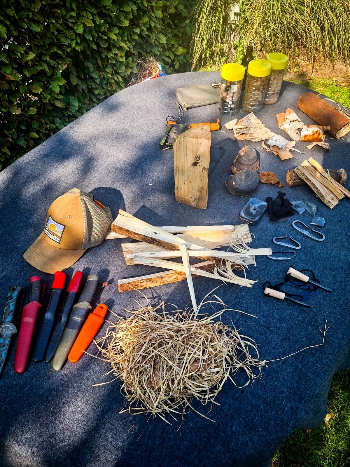 Outdoor table with camping gear, wood, and tools on a gray surface, surrounded by greenery.