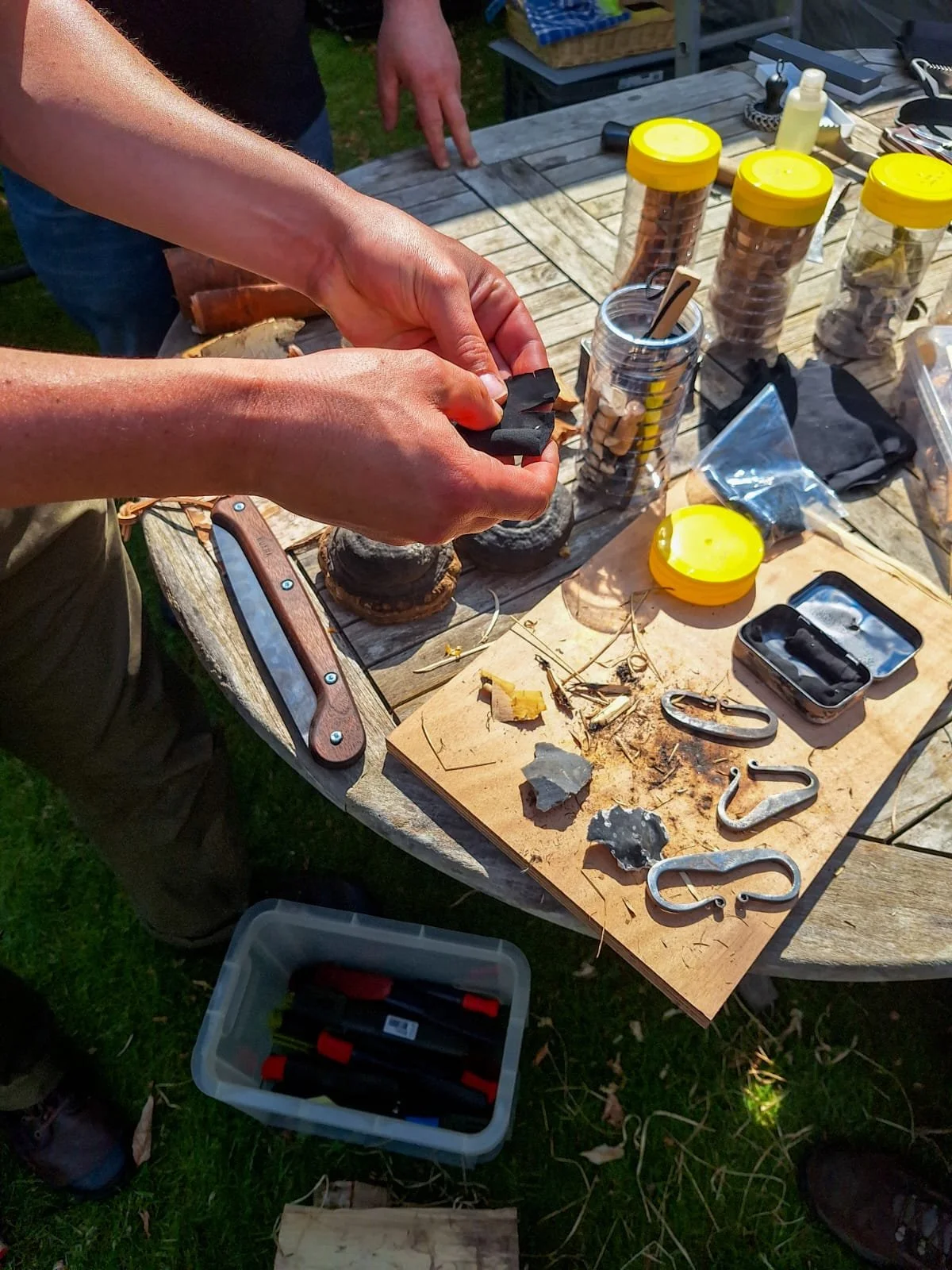 Person handling black flint or fire-starting material on a worn wooden cutting board with various metal hooks, rocks, and tools scattered on a weathered outdoor wooden table, surrounded by containers with fire-starting supplies and small items.
