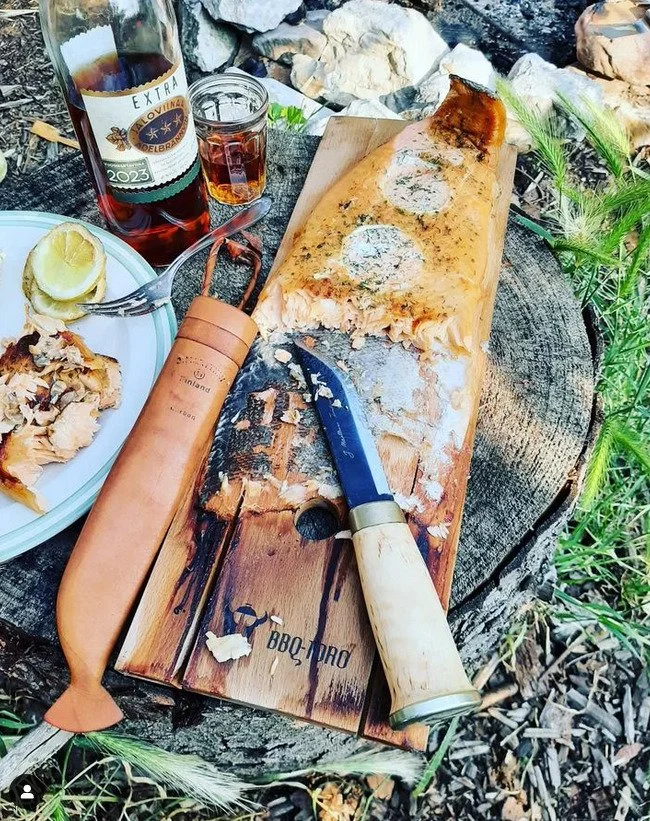 Outdoor picnic scene featuring a partially eaten pizza on a wooden board, a glass of iced tea or soda, a bottle of wine, a lemon wedge on a plate, and a meat tenderizer on a tree stump surrounded by grass and rocks.