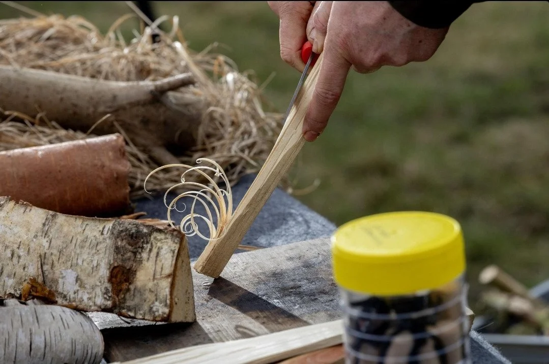A person using a wooden scraper to carve a piece of wood outdoors. Various wooden logs and sticks are on a table with a jar of nails or small metal objects nearby.