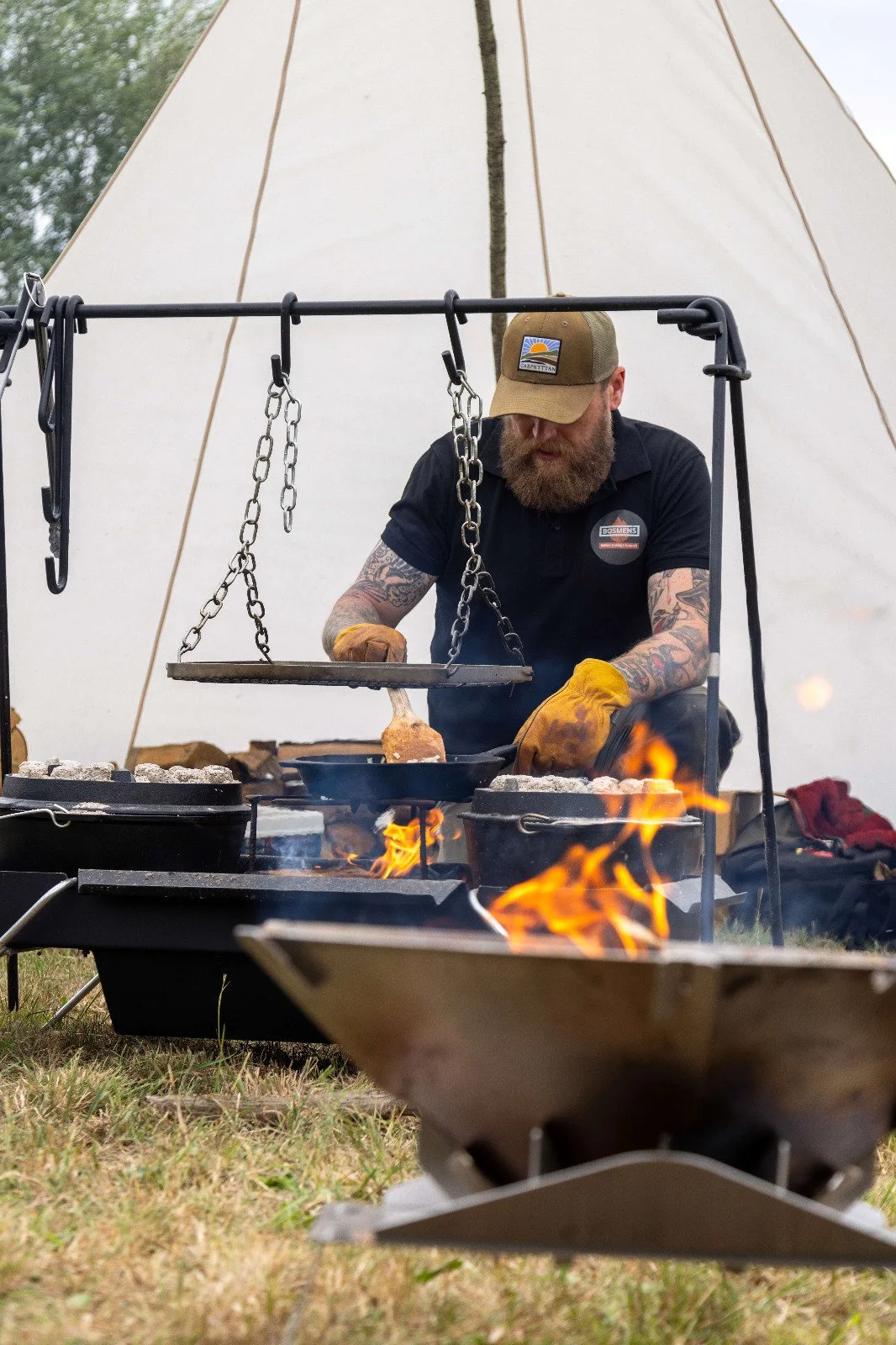 A man with tattoos and a beard wearing a tan cap and black shirt is cooking over an open flame. He is using a skillet to cook food outdoors, with metal chains hanging above the fire and a large beige tent in the background.