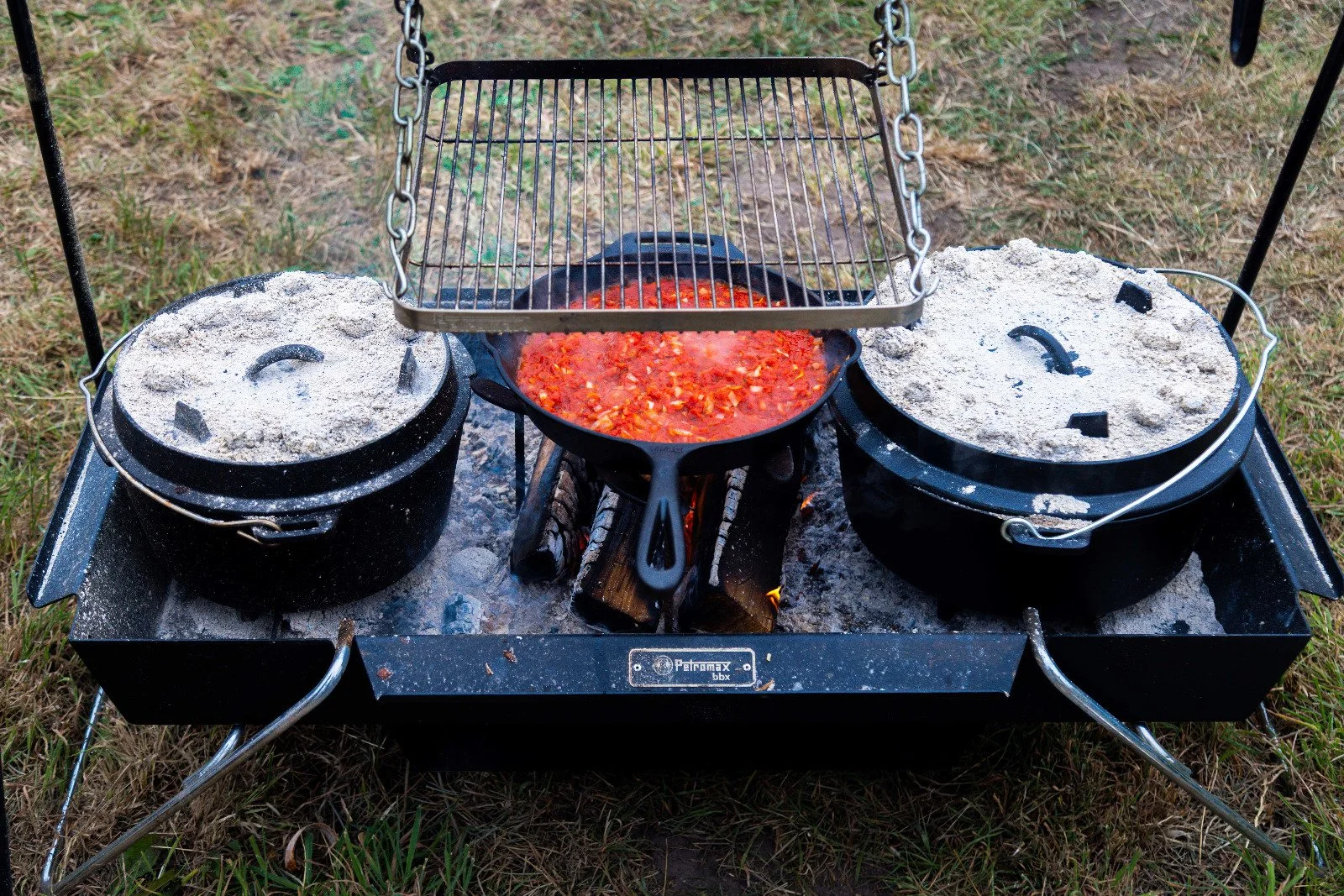 A camping stove with two covered pots on either side and a frying pan in the middle, cooking over an open flame on a grassy surface.