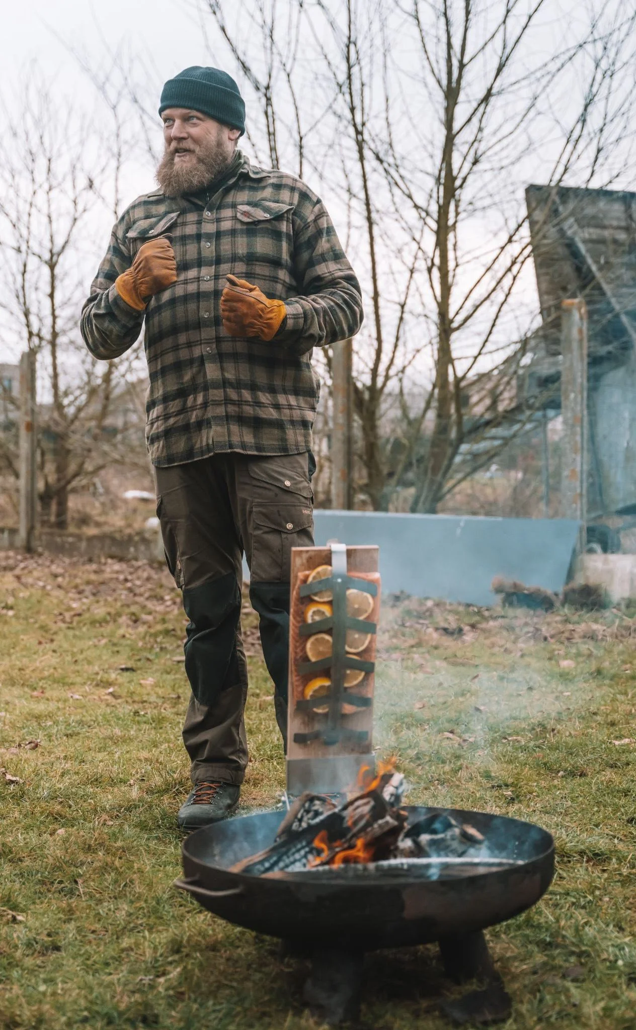 A man outdoors near a fire pit, wearing a beanie, plaid shirt, and gloves, standing with his fists near his chest, with a skewered food item above the fire.