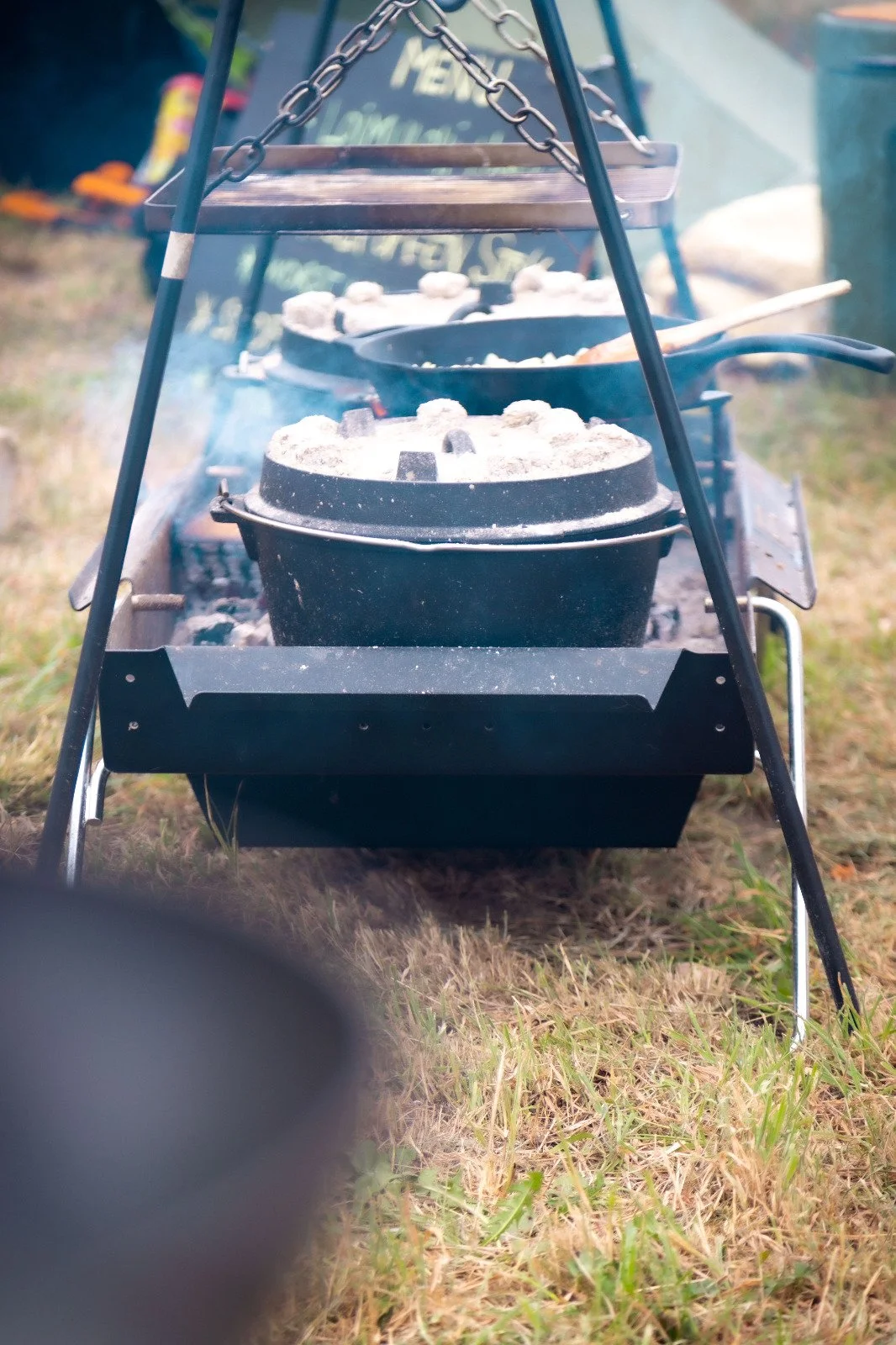 Outdoor camping scene with a portable grill, on which food is being cooked, and a frying pan filled with food, with grass on the ground.